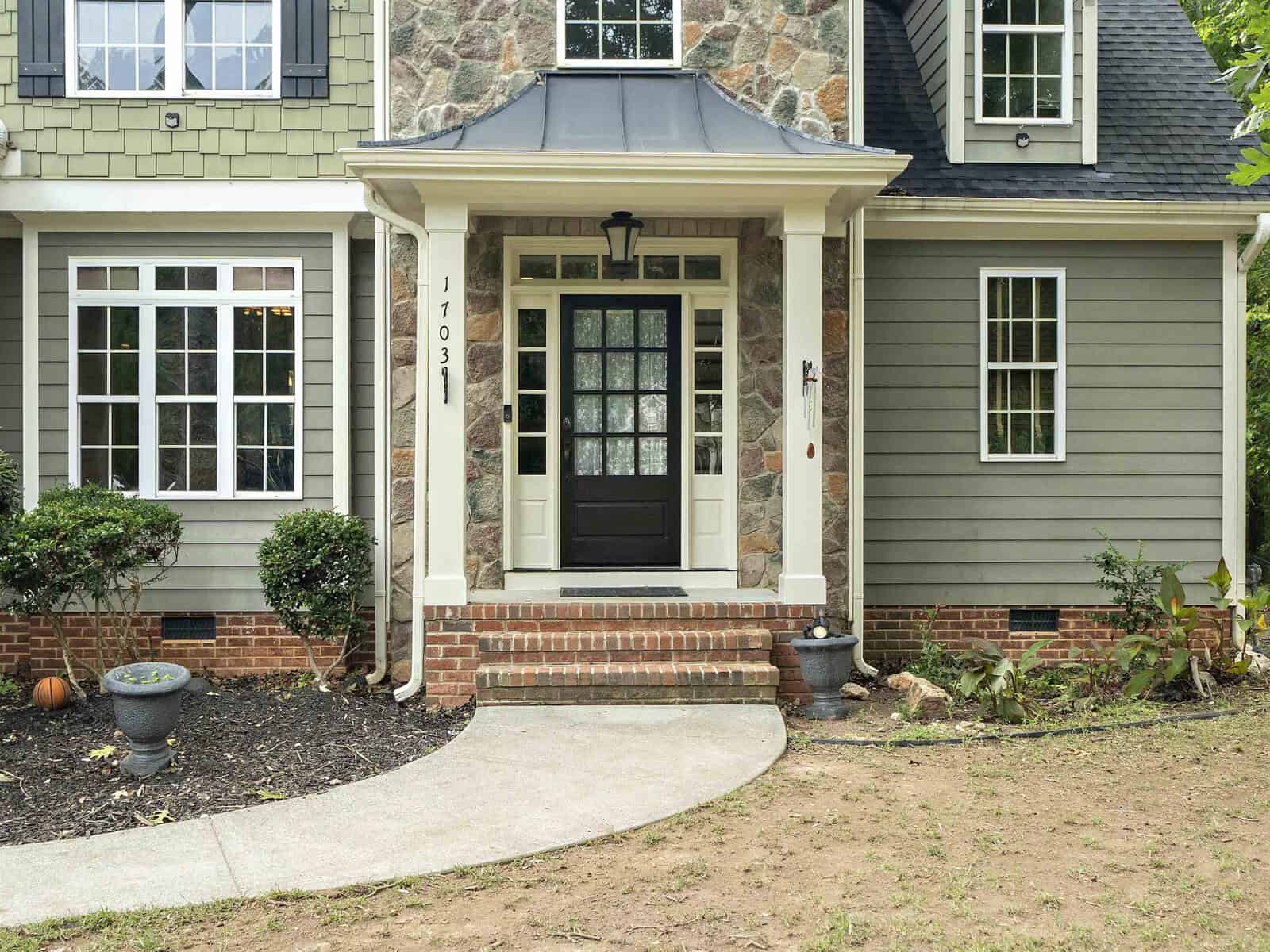 A two-story house with green siding and a stone facade. The front features a black door, framed windows, and a small porch with steps. There are shrubs and flower pots on either side of the entrance, and a curved walkway leading up to it.