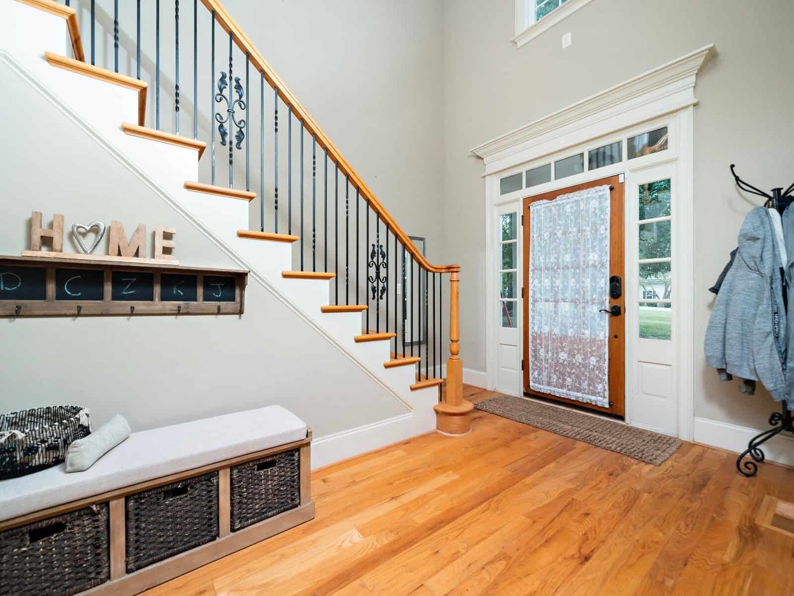 A bright, welcoming foyer featuring a staircase with black railings and wooden steps. A wooden bench with storage baskets beneath sits against the wall, adorned with home decor. A front door with lace curtains lets in natural light.