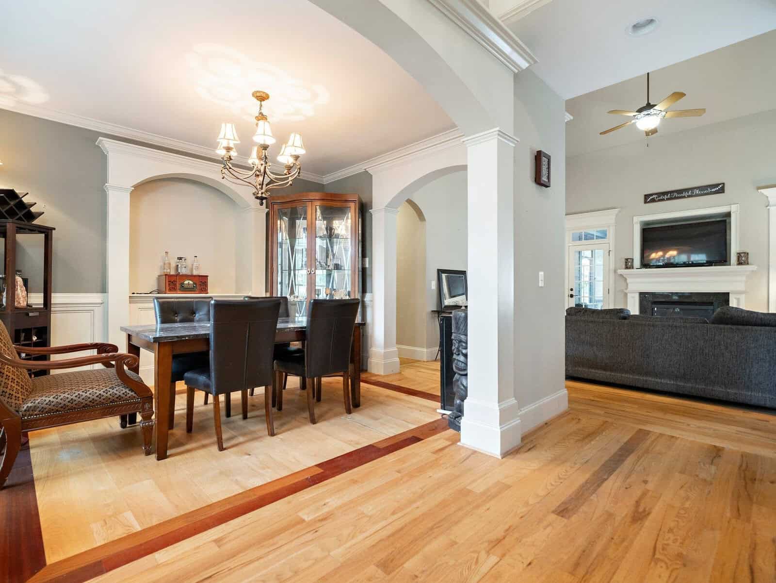 A dining room with a wooden table and black chairs next to a display cabinet. Adjacent, a living room with a gray couch faces a fireplace and TV. Arched doorways and light wood flooring connect the spaces. A chandelier and ceiling fan are visible.