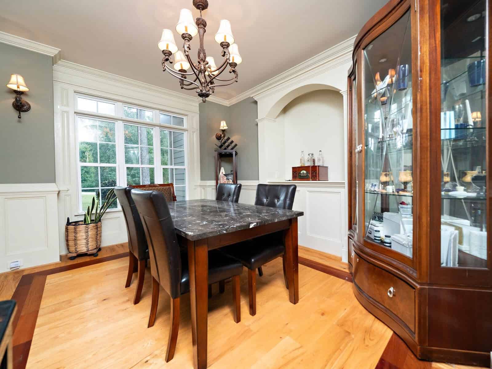 A cozy dining room with a wooden floor, featuring a dark marble table surrounded by four black leather chairs. A classic chandelier hangs above. A large window lets in natural light, and a china cabinet is in the corner displaying decorative items.