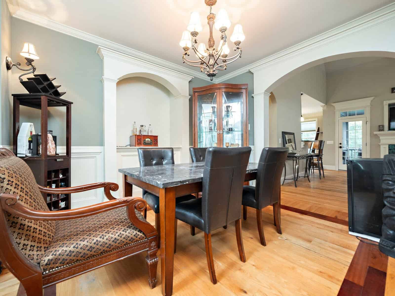 Spacious dining room featuring a wooden table with leather chairs, an ornate chandelier, decorative arches, and hardwood floors. Built-in shelves display various items, adding elegance to the warm, inviting atmosphere.