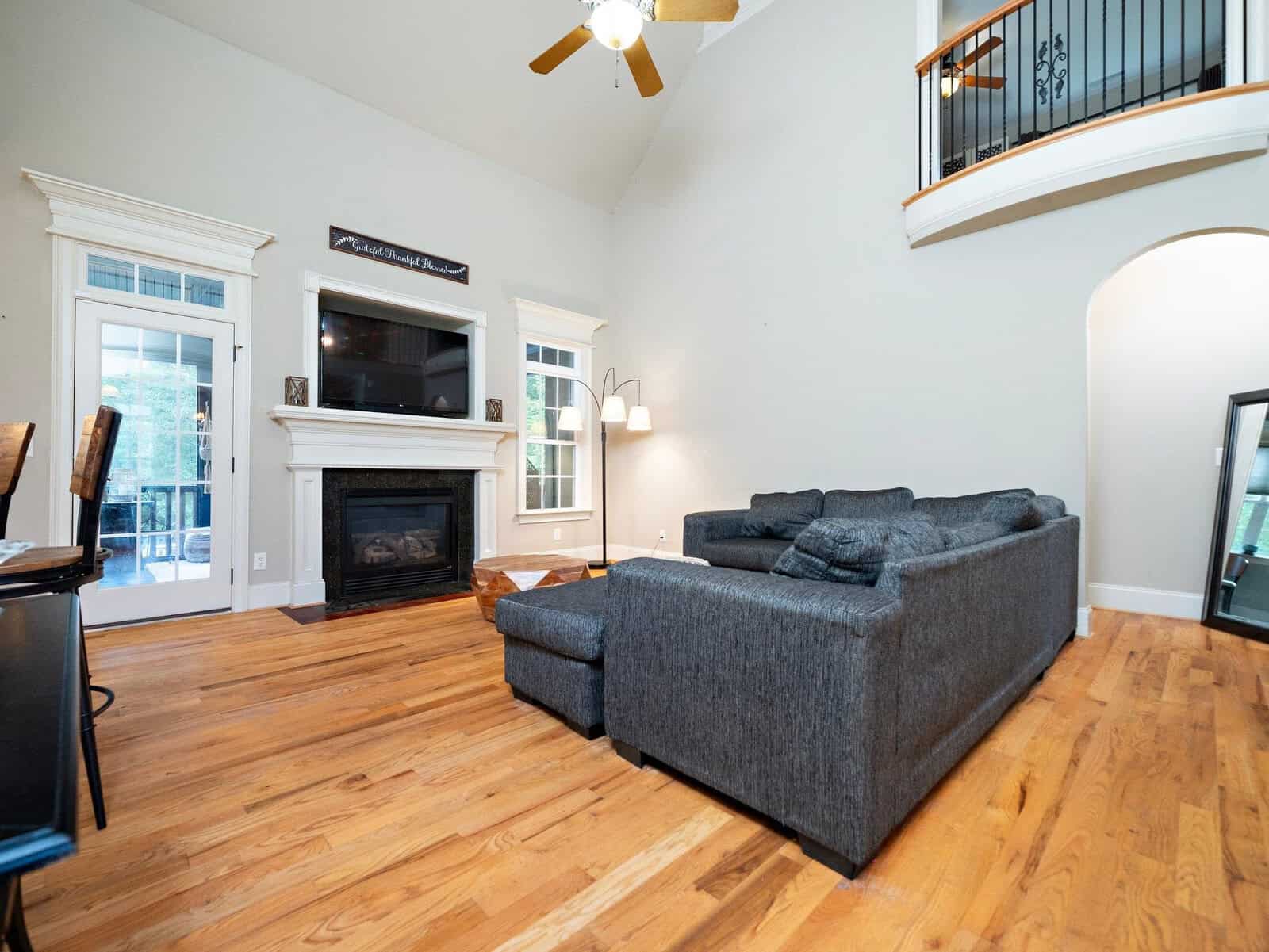 Spacious living room with high ceilings, featuring a gray sectional sofa, hardwood floors, and a fireplace with a TV above. A modern lamp stands beside the sofa, and a ceiling fan is positioned above. French doors and a curved balcony are visible.