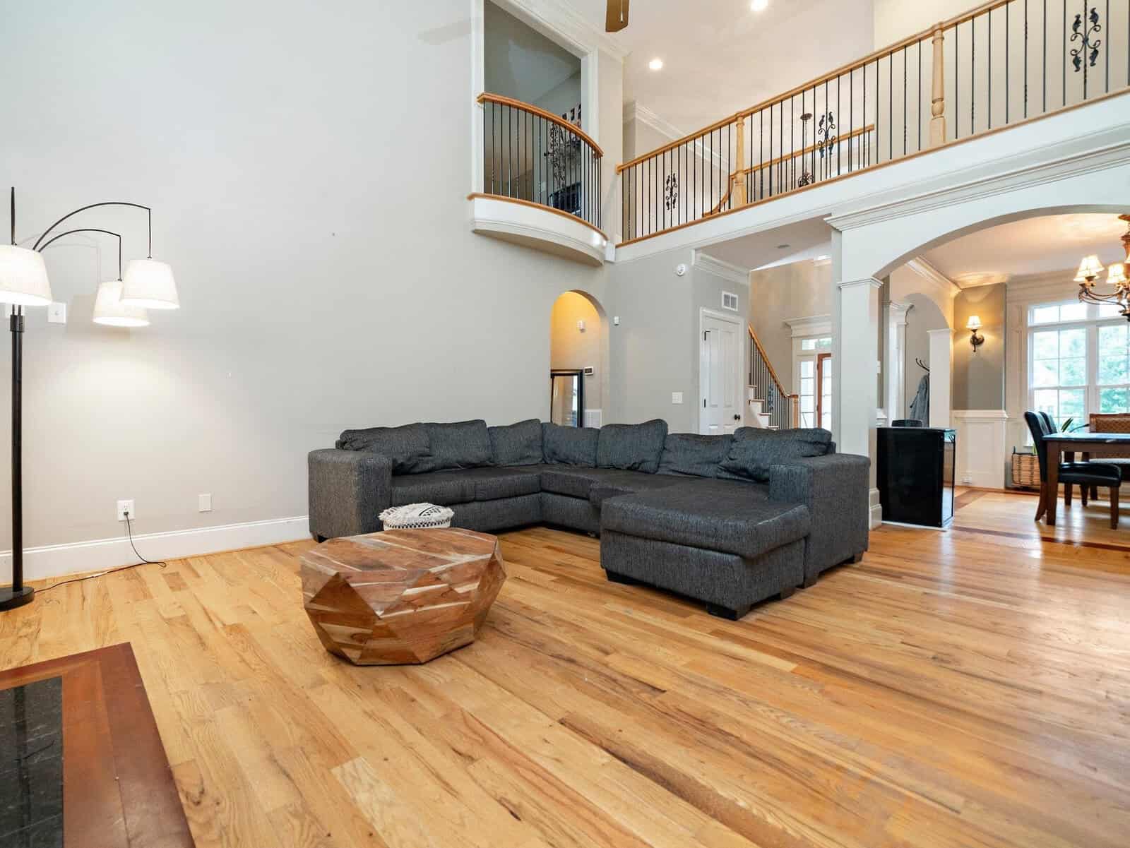 Spacious living room with a large gray sectional sofa, a wooden coffee table, and wooden floors. An open loft balcony overlooks the area. A dining room with a chandelier is visible in the background. The space is brightly lit by large windows.