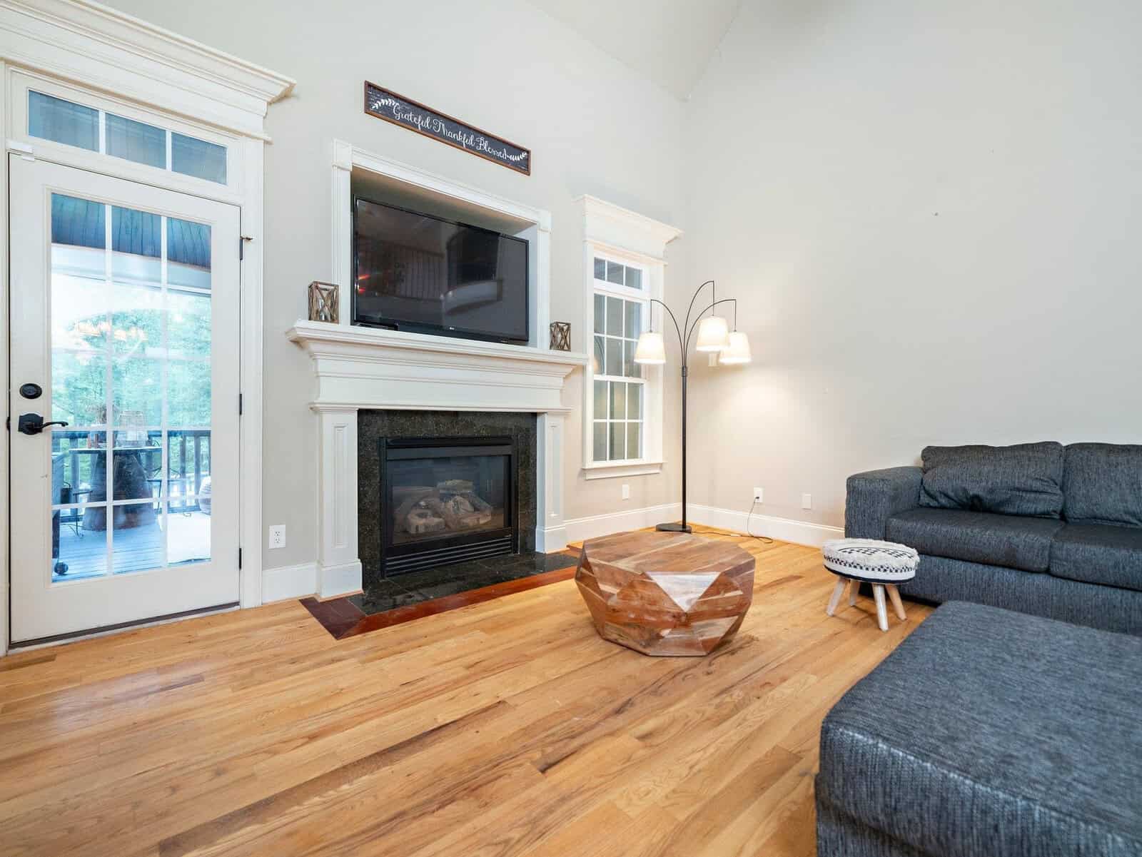 A cozy living room with a fireplace below a wall-mounted TV, a gray sectional sofa, and a modern floor lamp. There's a wooden coffee table on a hardwood floor, and to the left, a glass door leads to an outdoor patio.
