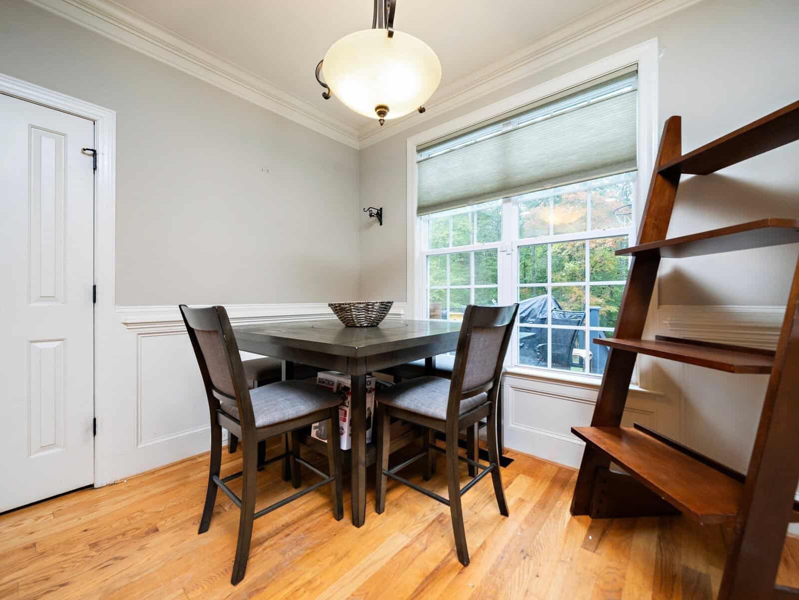 Dining room with a wooden table and four chairs. There's a wicker basket on the table. Large window with blinds on the right and a wooden ladder-style shelf in the corner. Light fixture hangs from the ceiling, and the floor is hardwood.