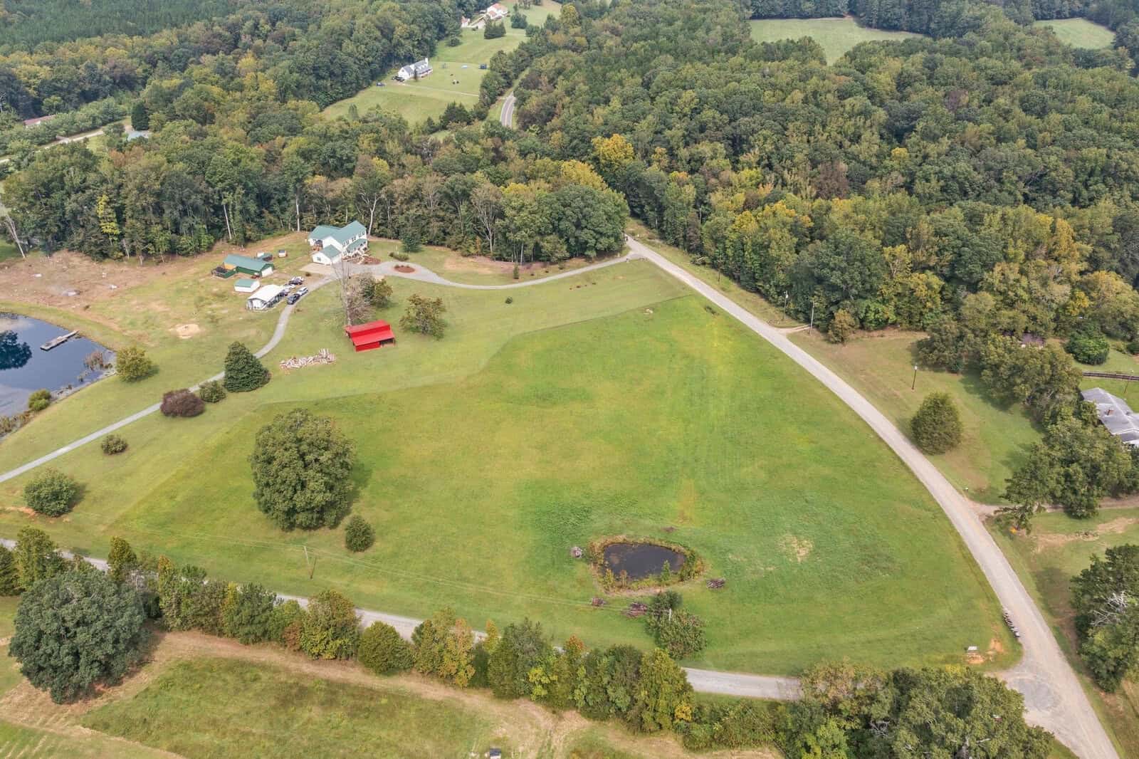An aerial view of Rougemont reveals a lush green landscape with a winding road leading to 5685 Hobgood Road. A small pond and red barn are nestled among the grassy areas, surrounded by dense woodlands and scattered buildings, crafting a picturesque rural scene.