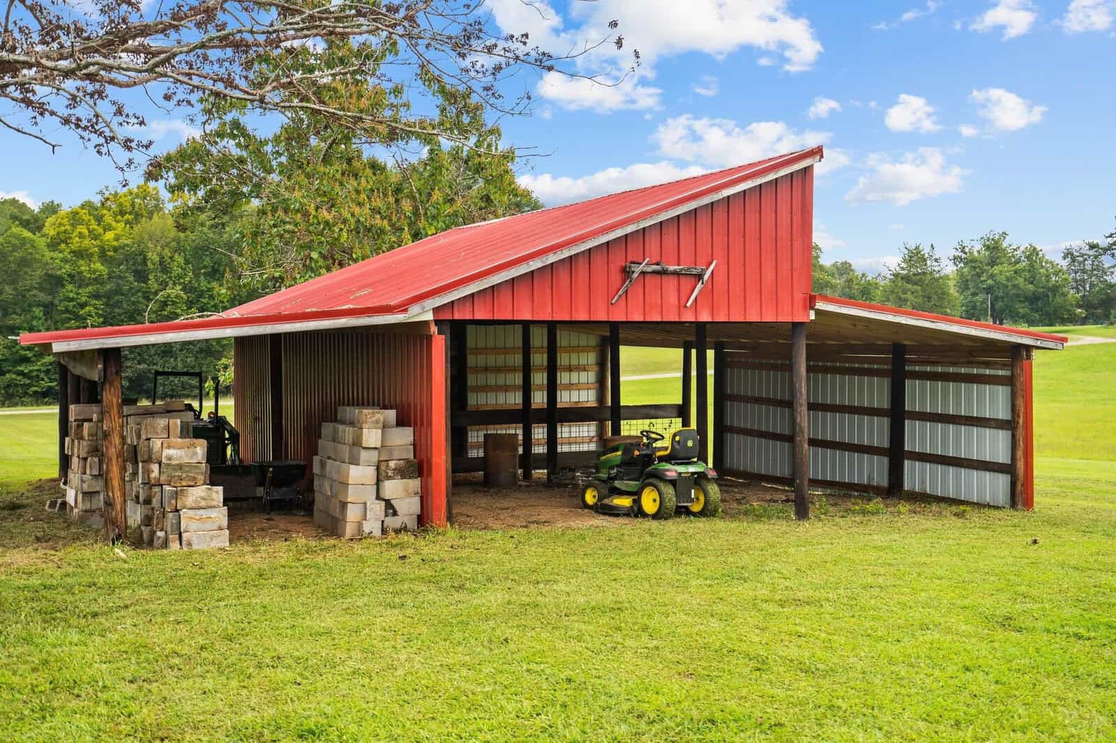 A red-roofed shed stands in a grassy field under a partly cloudy sky. The shed houses a green and yellow riding mower and stacks of large bricks. Trees and a gently sloping hill are visible in the background.
