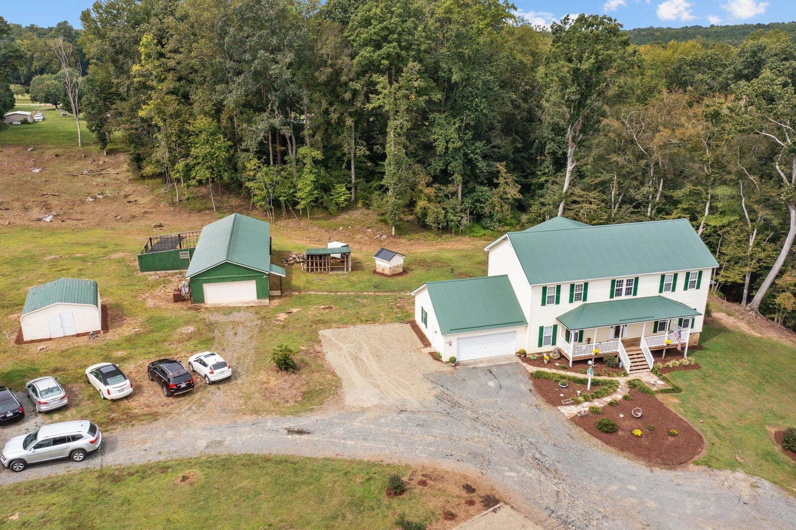 Aerial view of a rural property with a two-story house featuring a green roof. Nearby are a couple of smaller structures, parked cars, and a gravel driveway. The area is surrounded by trees and an open field.