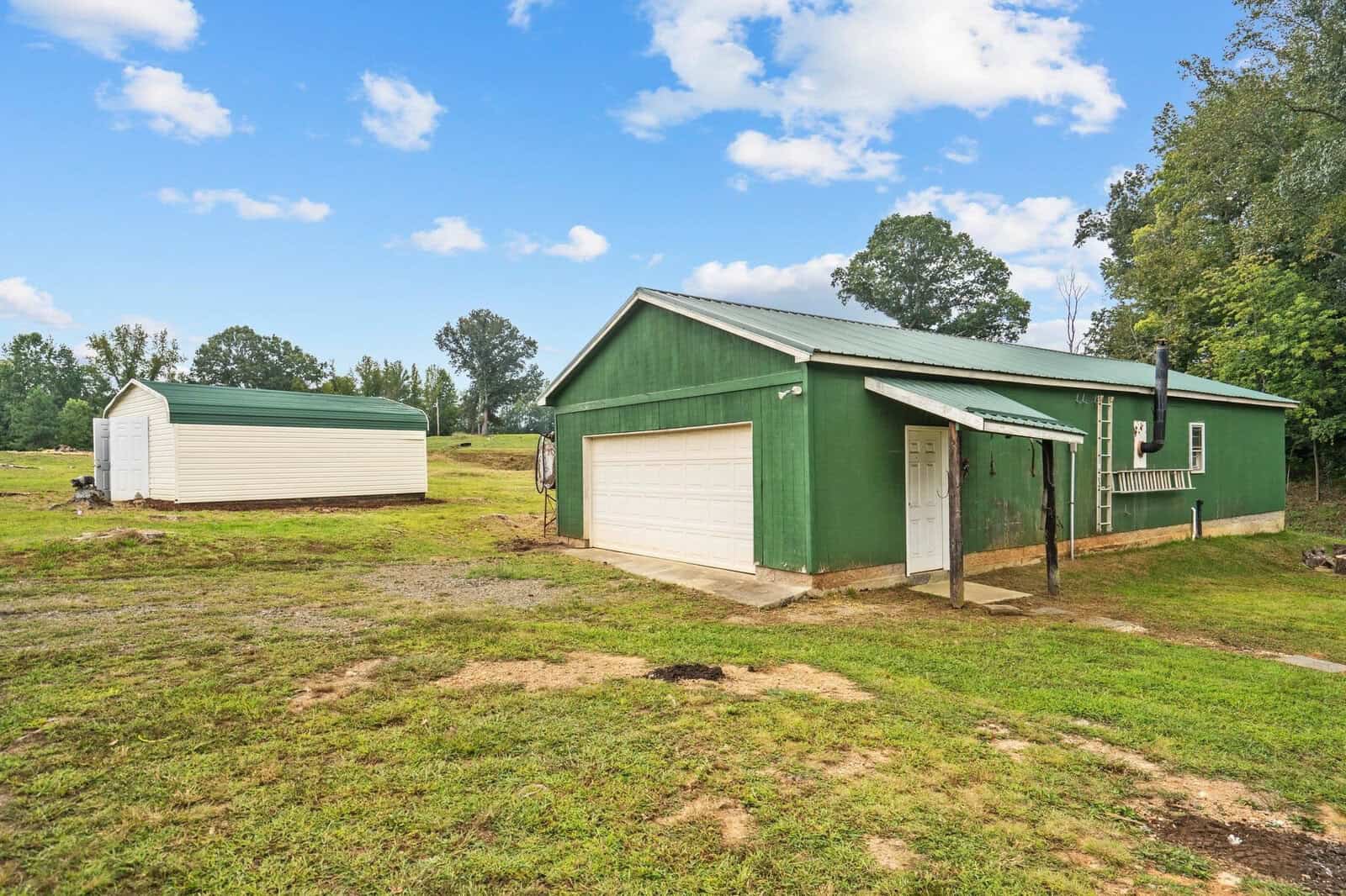 A green building with a white garage next to a smaller white shed in a grassy yard. The sky is blue with scattered clouds, and there are trees in the background.