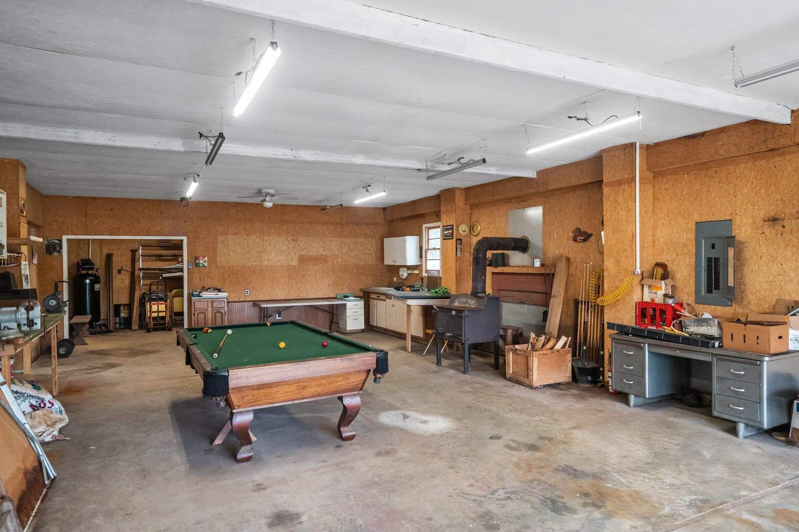 A spacious garage with a pool table at the center, surrounded by workbenches, tools, and storage shelves. The room has a concrete floor and wooden walls, with fluorescent lighting overhead and various items scattered around.