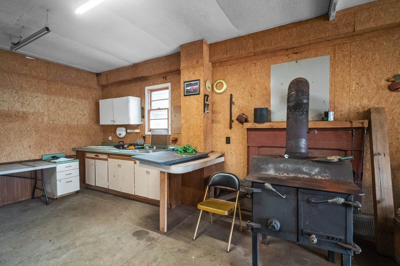 A rustic workshop with wooden walls features a wood-burning stove and a small kitchen area. The space includes cabinets, a counter with various tools, a chair, and wall decorations, creating a cozy, utilitarian atmosphere.