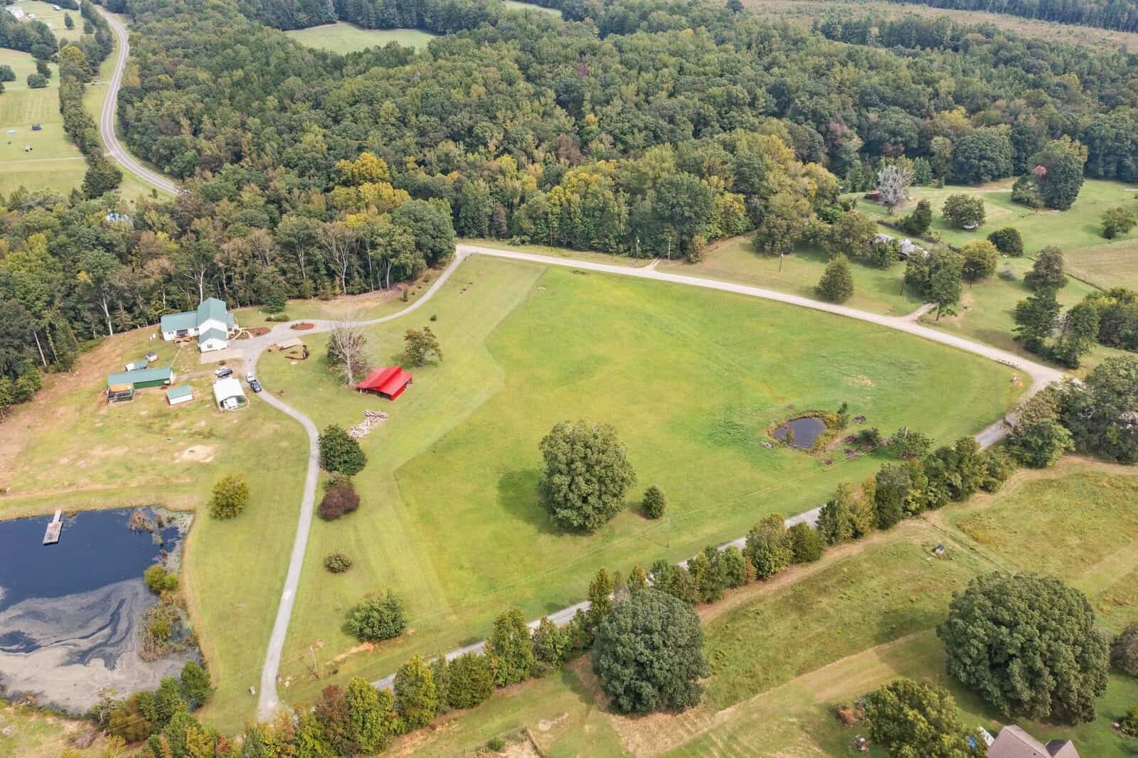 Aerial view of a rural landscape with a large grassy area, a winding road, a pond, and several buildings. Trees surround the area, and a red structure is visible near the center. Woods extend into the distance.