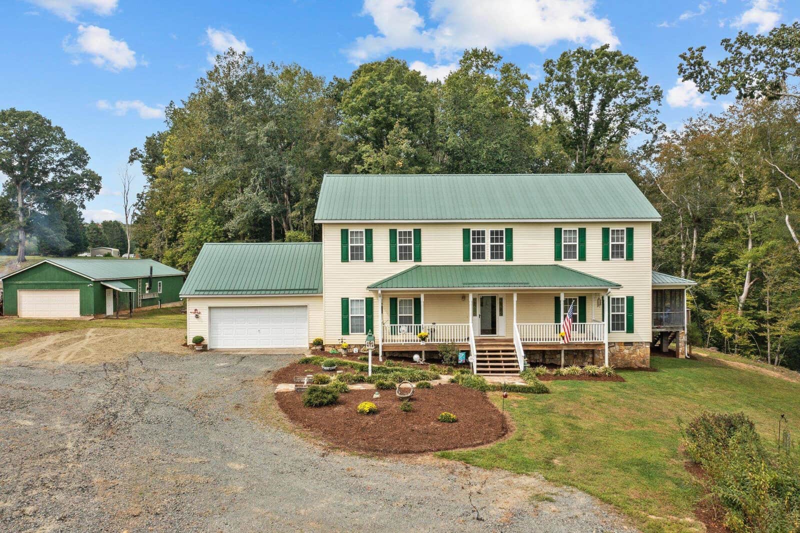 A two-story house with a green metal roof, white siding, and green shutters, surrounded by trees. A well-maintained front yard has a small garden. A gravel driveway leads to the garage. A separate green-roofed building is visible nearby.
