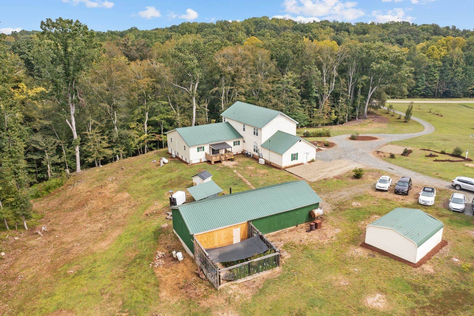 Aerial view of a rural property featuring a large white house with a green roof, multiple outbuildings, and several cars parked nearby. The area is surrounded by trees and a grassy field, providing a secluded and peaceful setting.