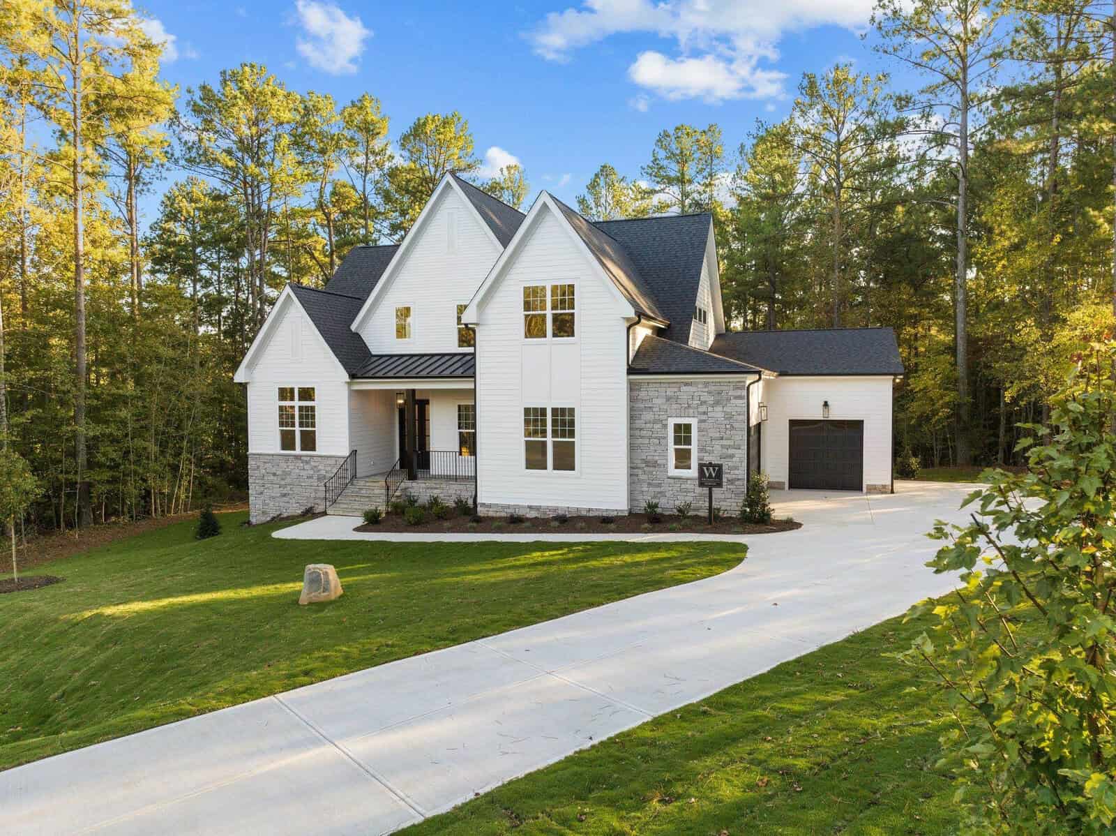 Located at 3912 Sage Court, this expansive two-story white house in Wake Forest boasts stone accents, a black roof, and a spacious driveway. It is surrounded by a manicured lawn and tall trees under a clear blue sky.