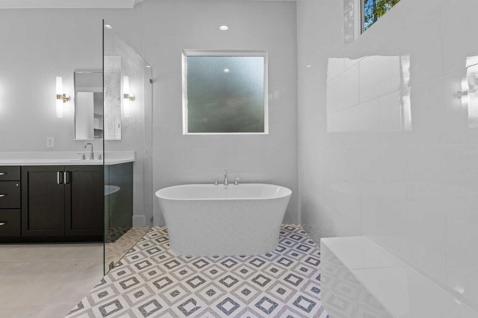 A modern bathroom featuring a freestanding bathtub on a patterned tile floor. A large frosted window provides natural light. A glass shower enclosure is on one side, with a double sink vanity having dark cabinetry on the other.