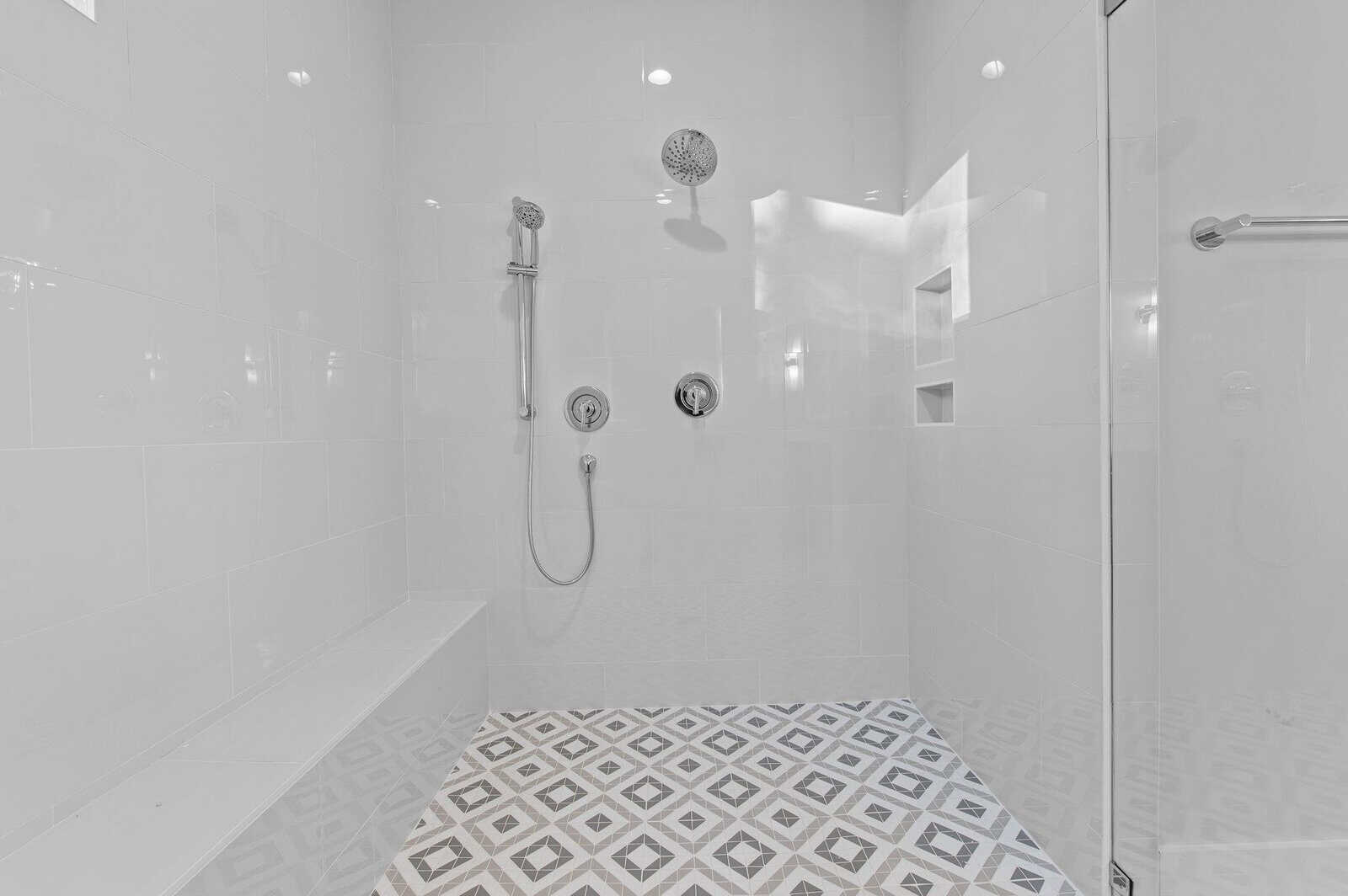 A modern, bright white tiled shower with geometric patterned floor tiles. It features a wall-mounted showerhead, a handheld shower, built-in shelves, and a bench. The shower area is enclosed by glass doors.