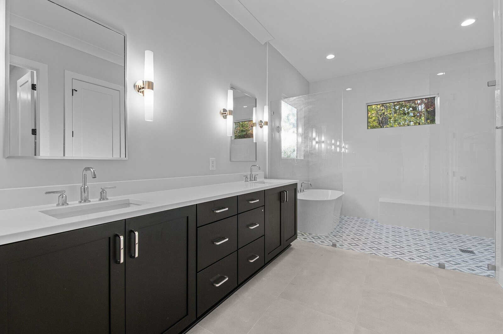 A modern bathroom with a double-sink vanity with black cabinets and white countertops. There's a large mirror above the sinks, a freestanding bathtub, and a walk-in shower with a small window. The floor features a geometric tile pattern.