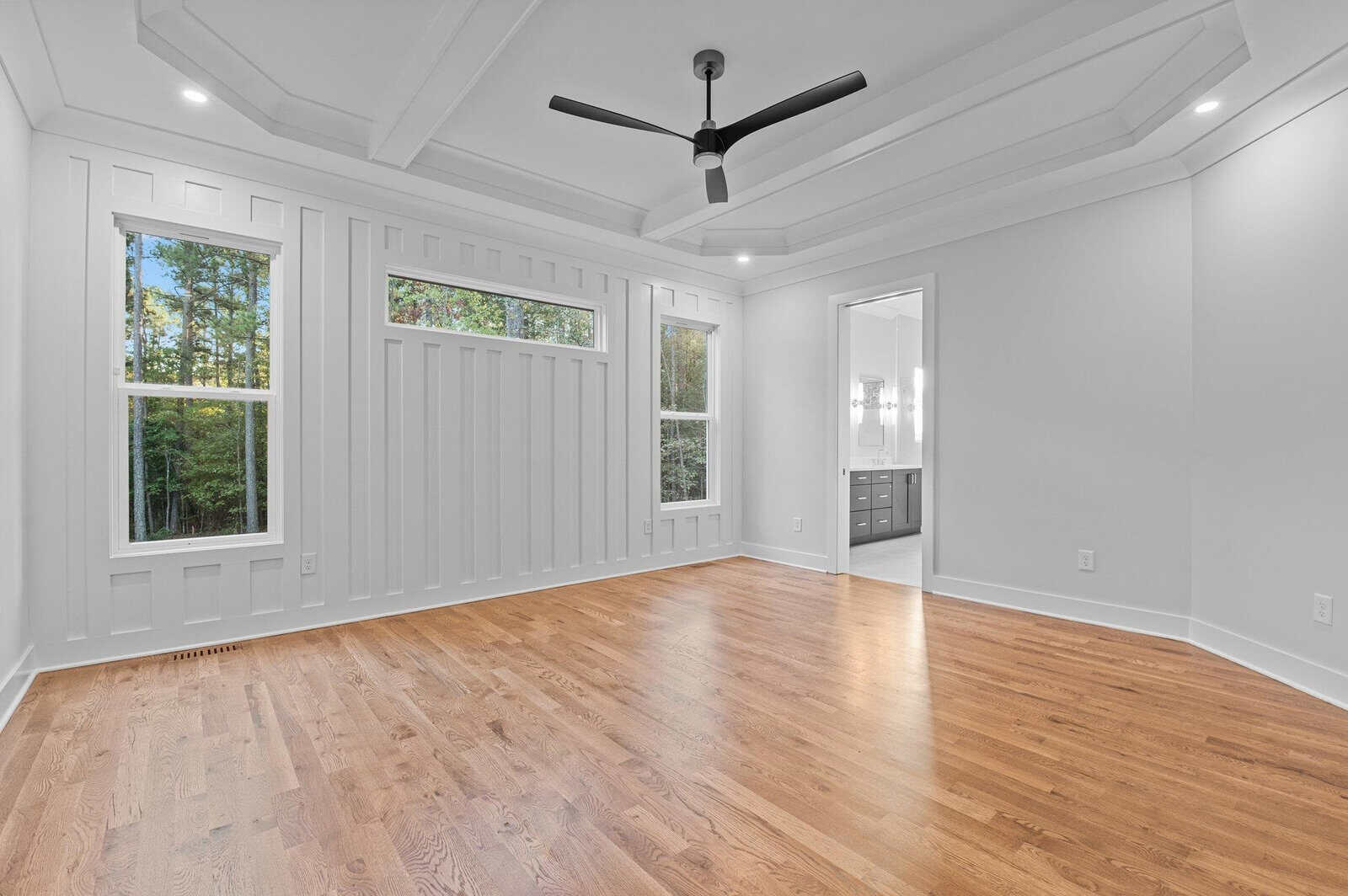 A spacious empty room with light wooden floors, white walls, and large windows showing a view of trees. A black ceiling fan hangs from the coffered ceiling. An open doorway leads to a bathroom with a double vanity.