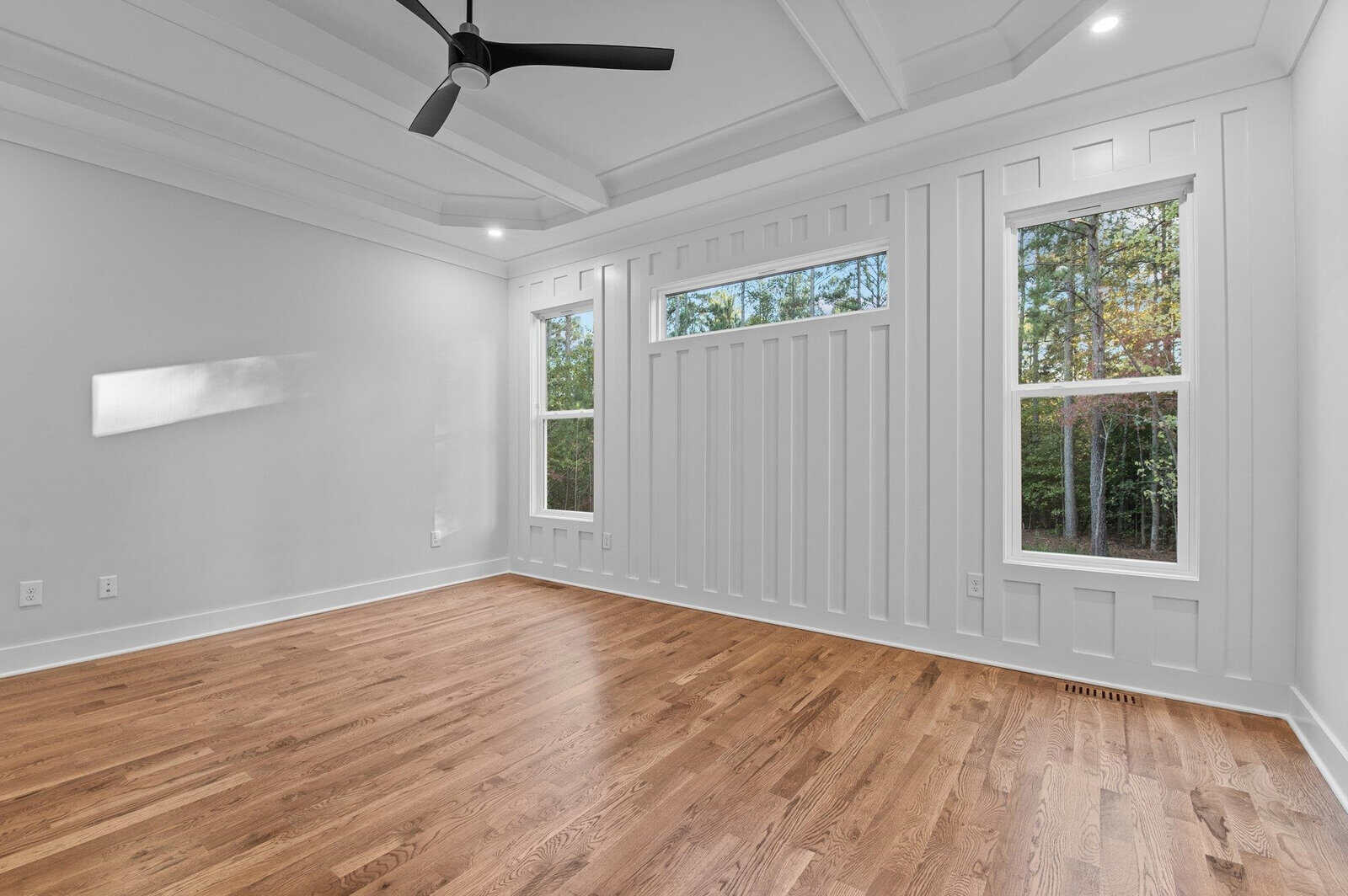 Empty room with light wooden floors, white paneled walls, and two tall windows framing a view of greenery outside. Ceiling features a fan and decorative molding. Natural light illuminates the space.