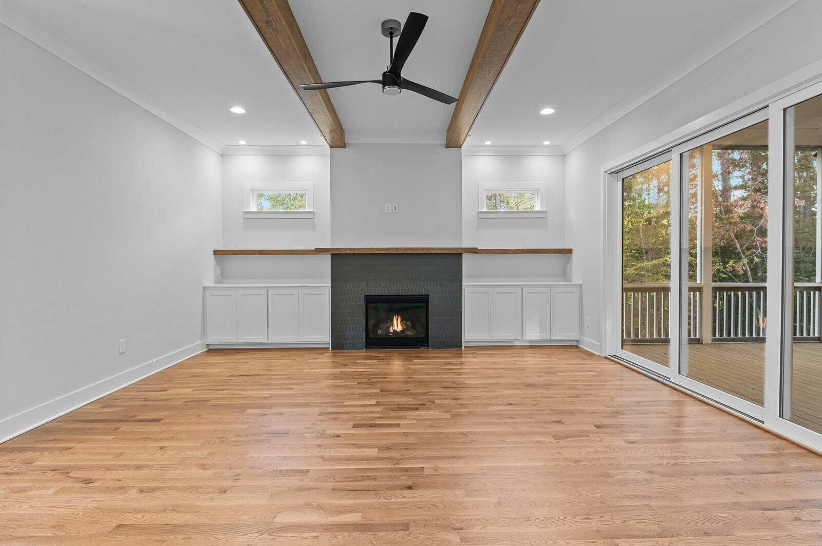 A modern living room with hardwood floors and a black fireplace centered on a wall featuring wood beams. Two small windows flank the fireplace. Large sliding glass doors lead to an outdoor deck with trees visible outside.