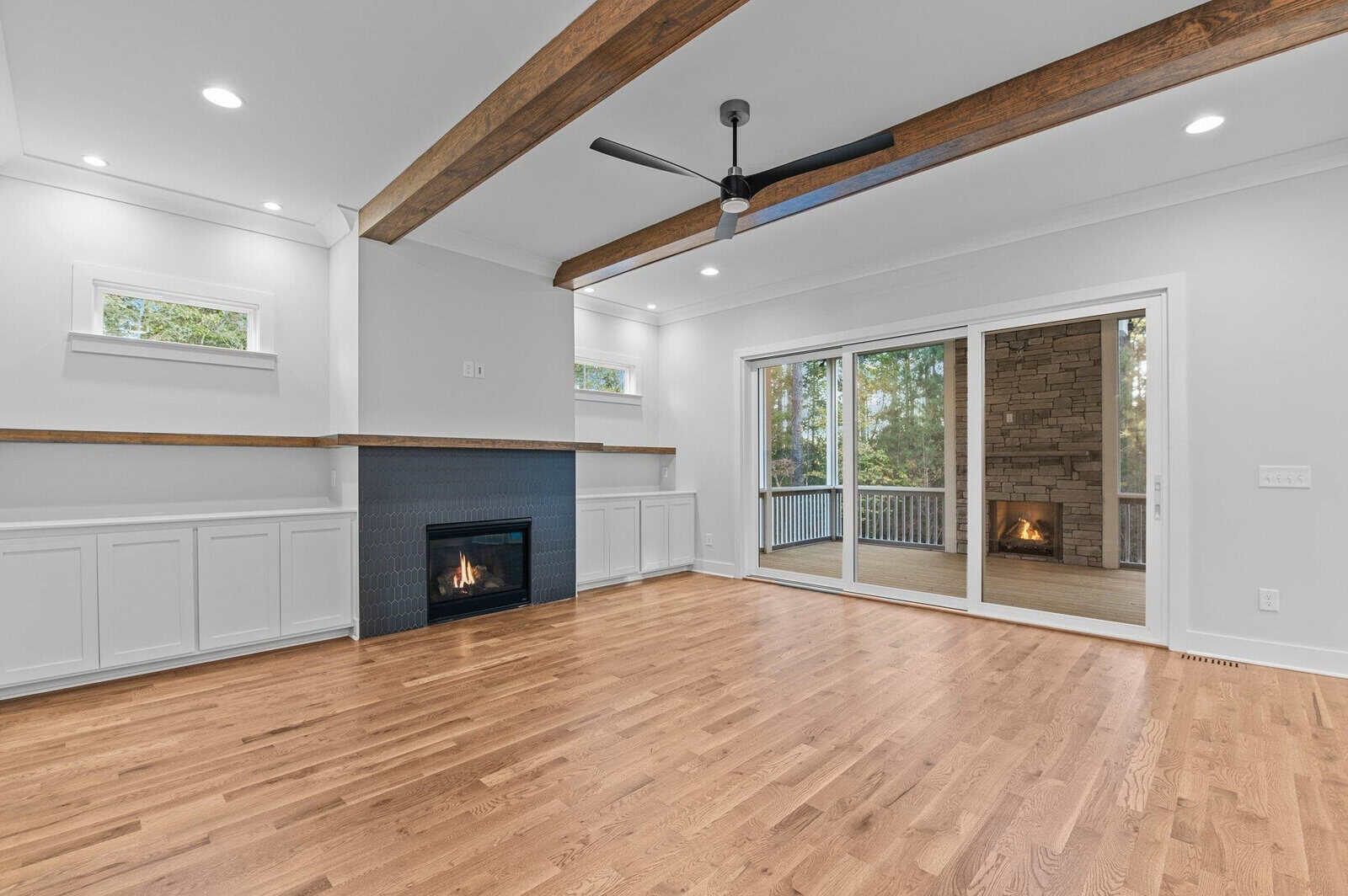 A bright living room with light wood floors, a black fireplace, and white built-in cabinets. Exposed wooden ceiling beams and a central fan add character. Large glass doors open to a screened patio with a brick fireplace and tree views.