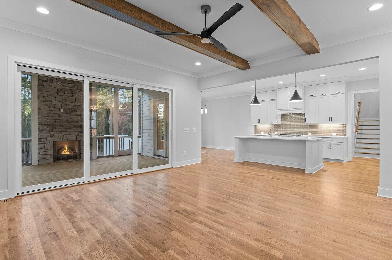 Spacious modern living area with wooden floors, exposed wooden ceiling beams, and a ceiling fan. Features a large kitchen with white cabinets and pendant lights, and a view of an outdoor patio with a fireplace through sliding glass doors.