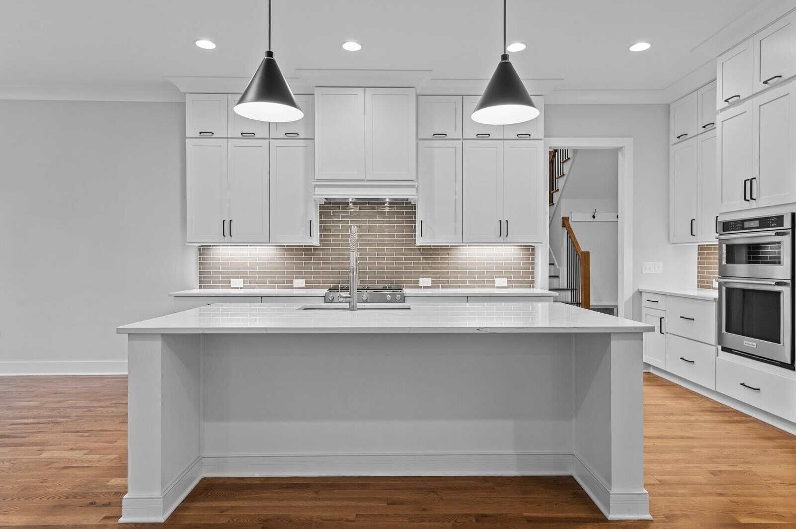 A modern kitchen with white cabinets and a central island topped with a white countertop. Two black pendant lights hang above the island. The backdrop features a glossy tiled backsplash and stainless steel appliances. The floor is wooden.