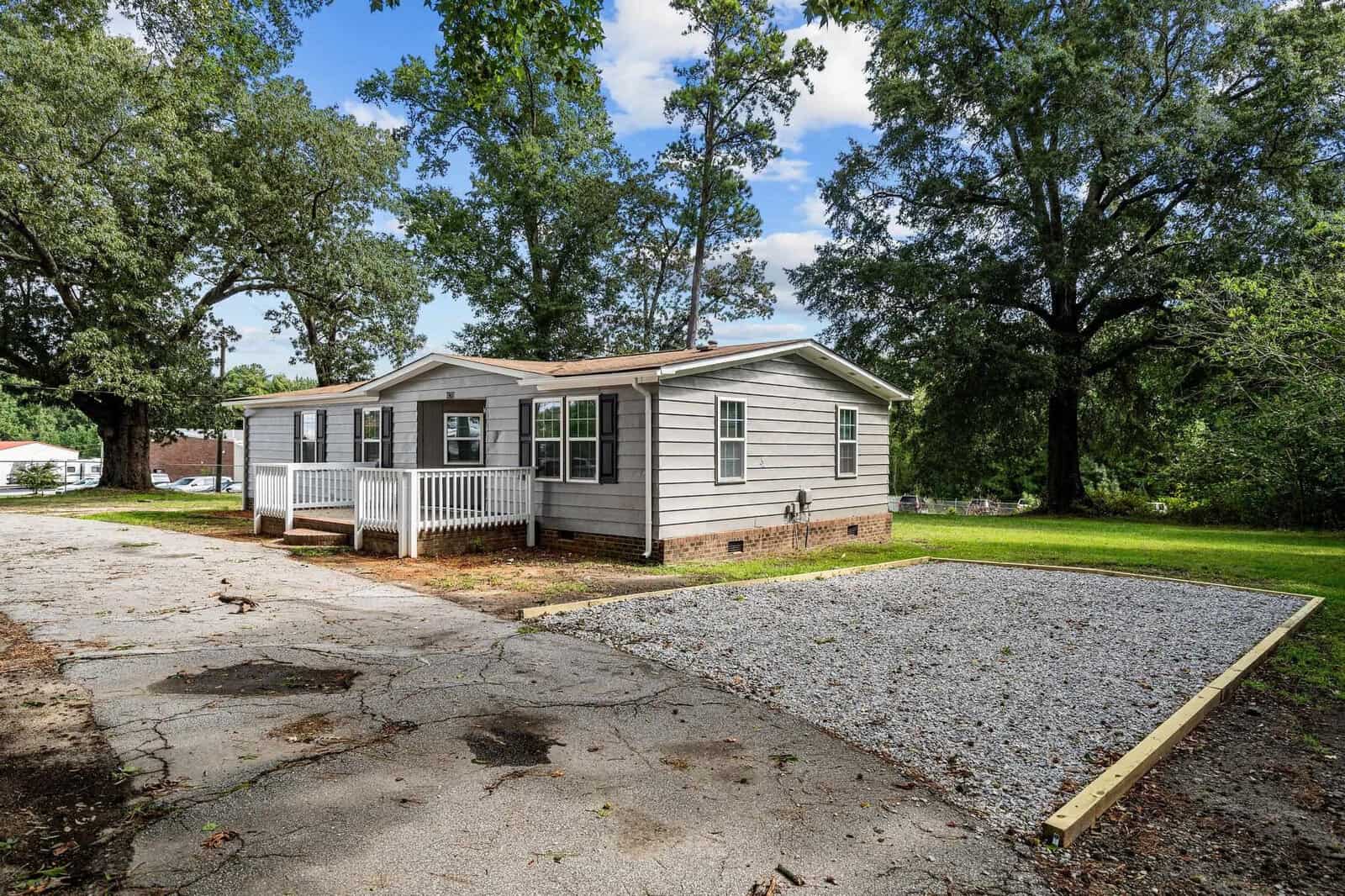 A single-story modular home with a small front porch, surrounded by trees and a partly paved driveway in the foreground.