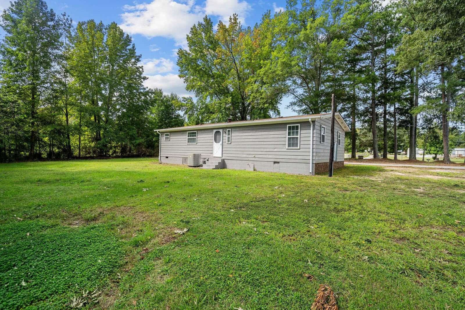 A single-story gray mobile home located in a grassy area with tall trees in the background on a sunny day.