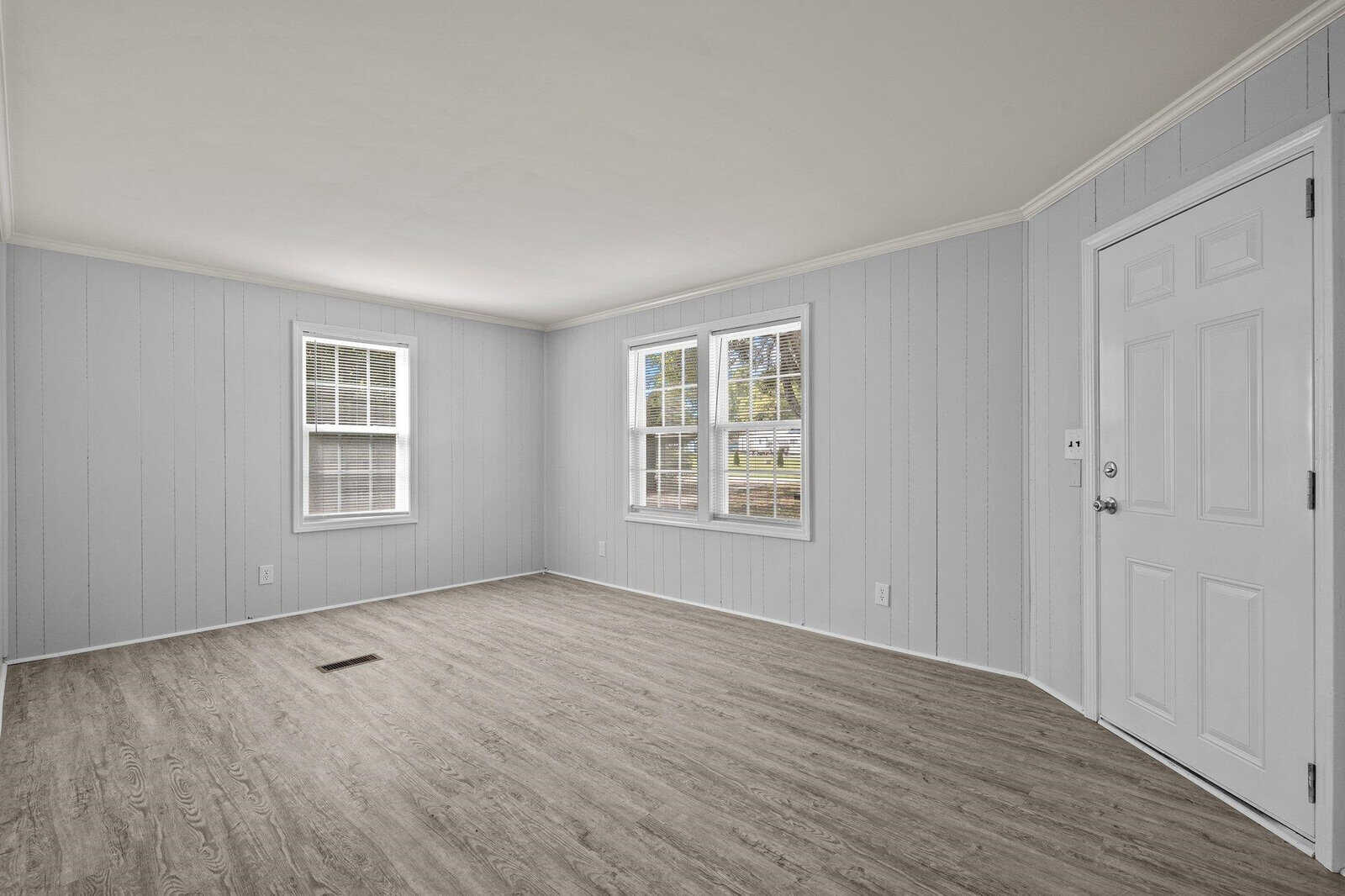 Empty room with light grey walls, two windows with white blinds, and wood laminate flooring. a white door is situated on the right side of the room.