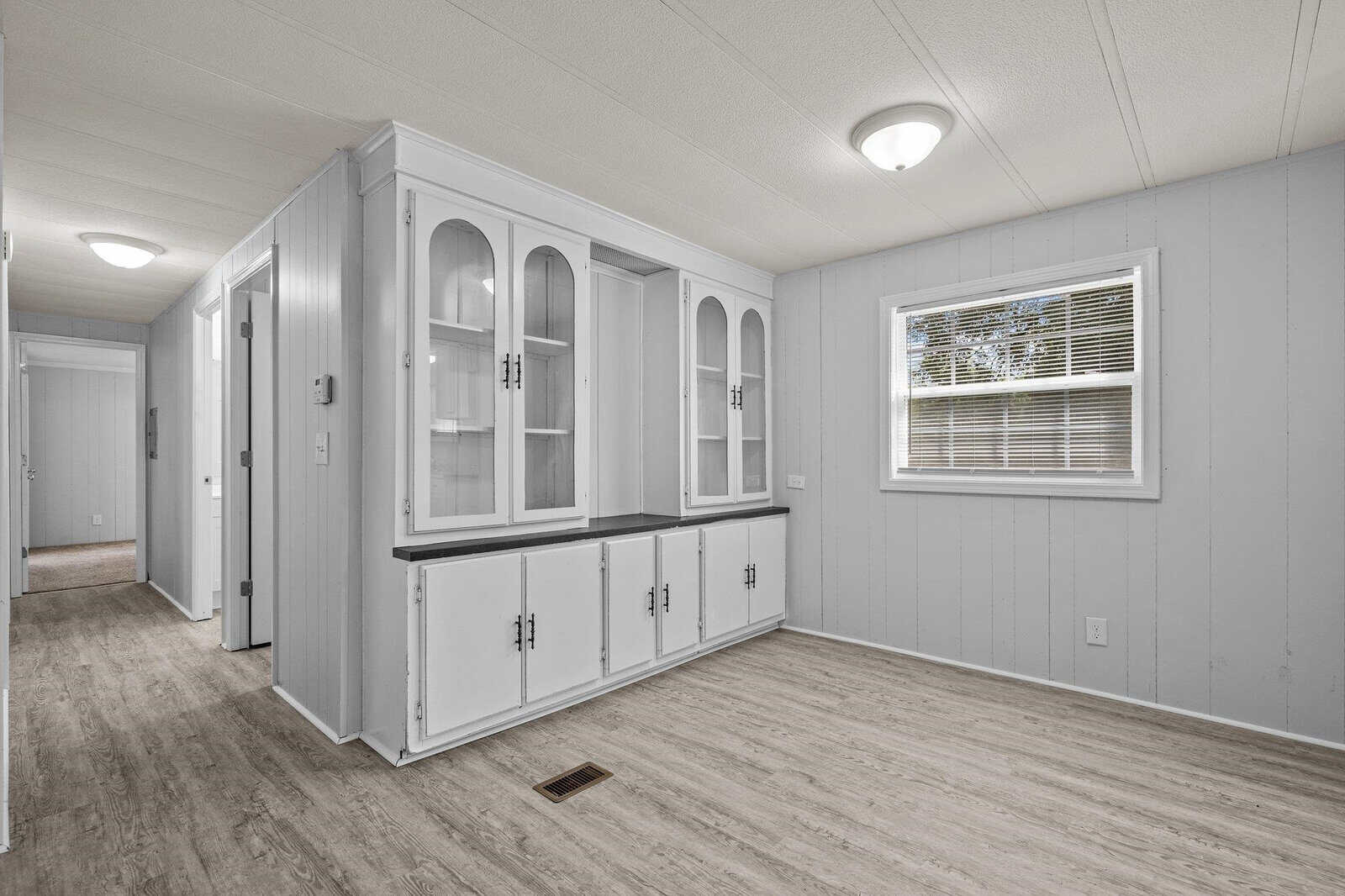 A bright, empty room featuring pale gray walls, white cabinetry with glass doors, and light wood laminate flooring. a window with closed blinds is present on the right side wall.