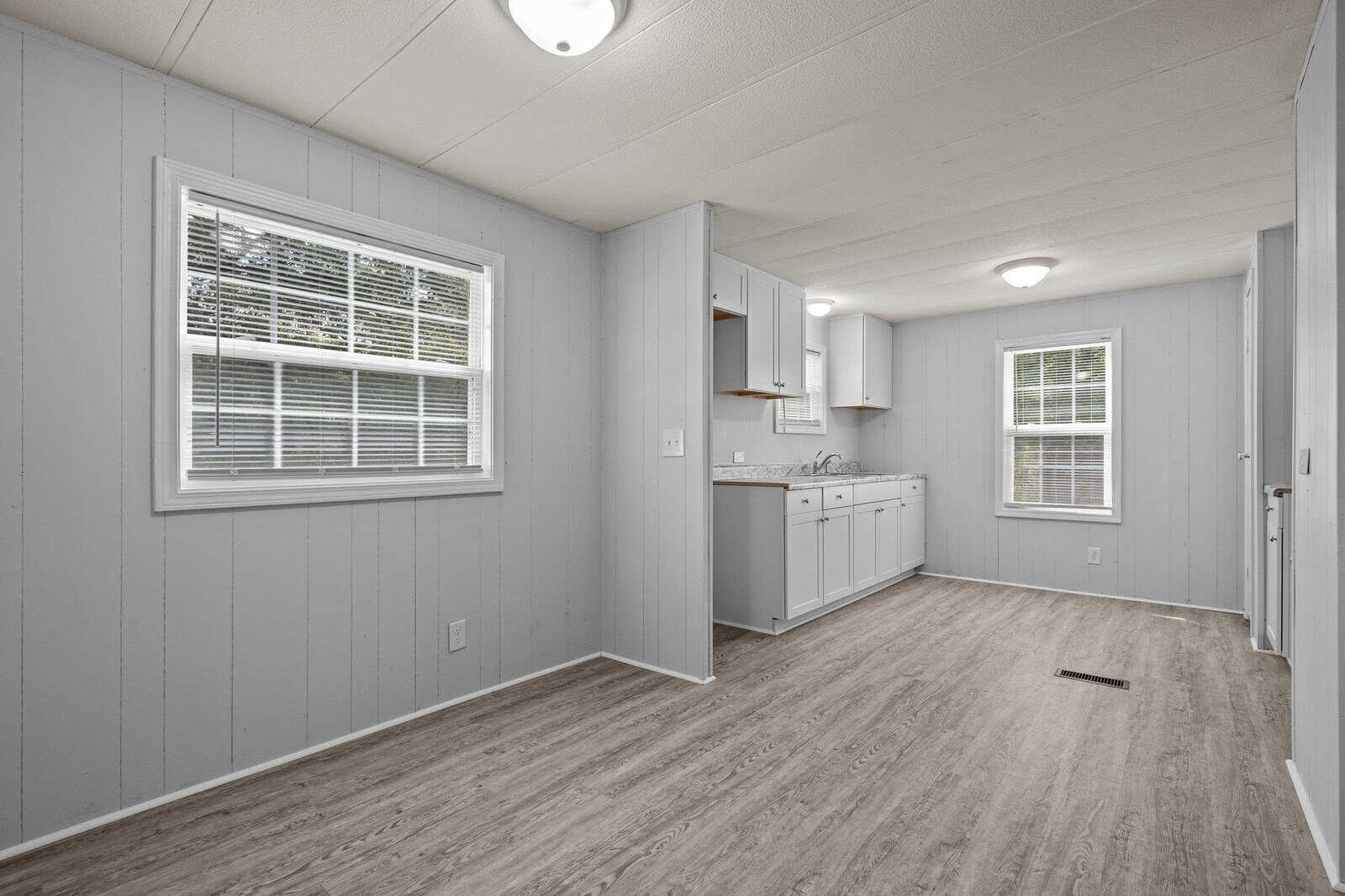 A small, empty kitchen and hallway with gray walls, white trim, wood laminate flooring, and two windows with closed horizontal blinds.