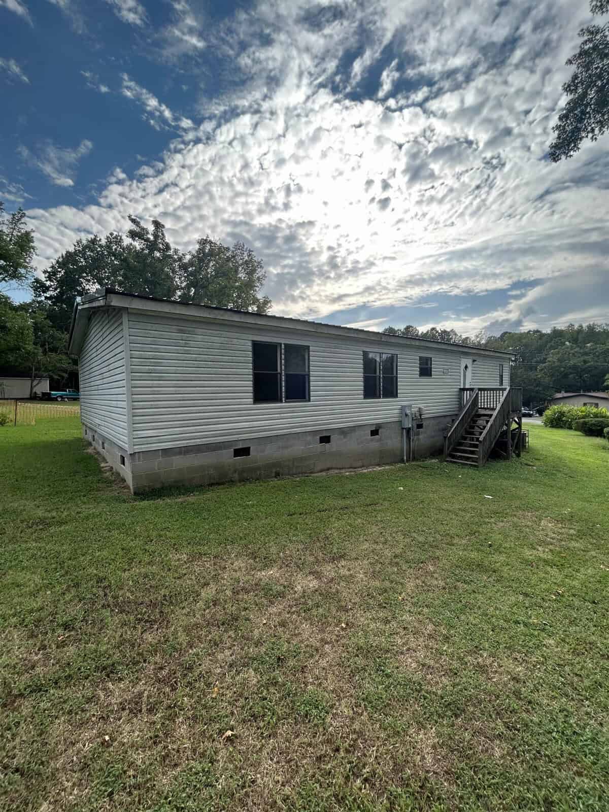 A large mobile home with white siding is set on a grassy lawn. A staircase leads to the entrance on the right. The sky is partly cloudy with patches of blue, and trees are visible in the background.