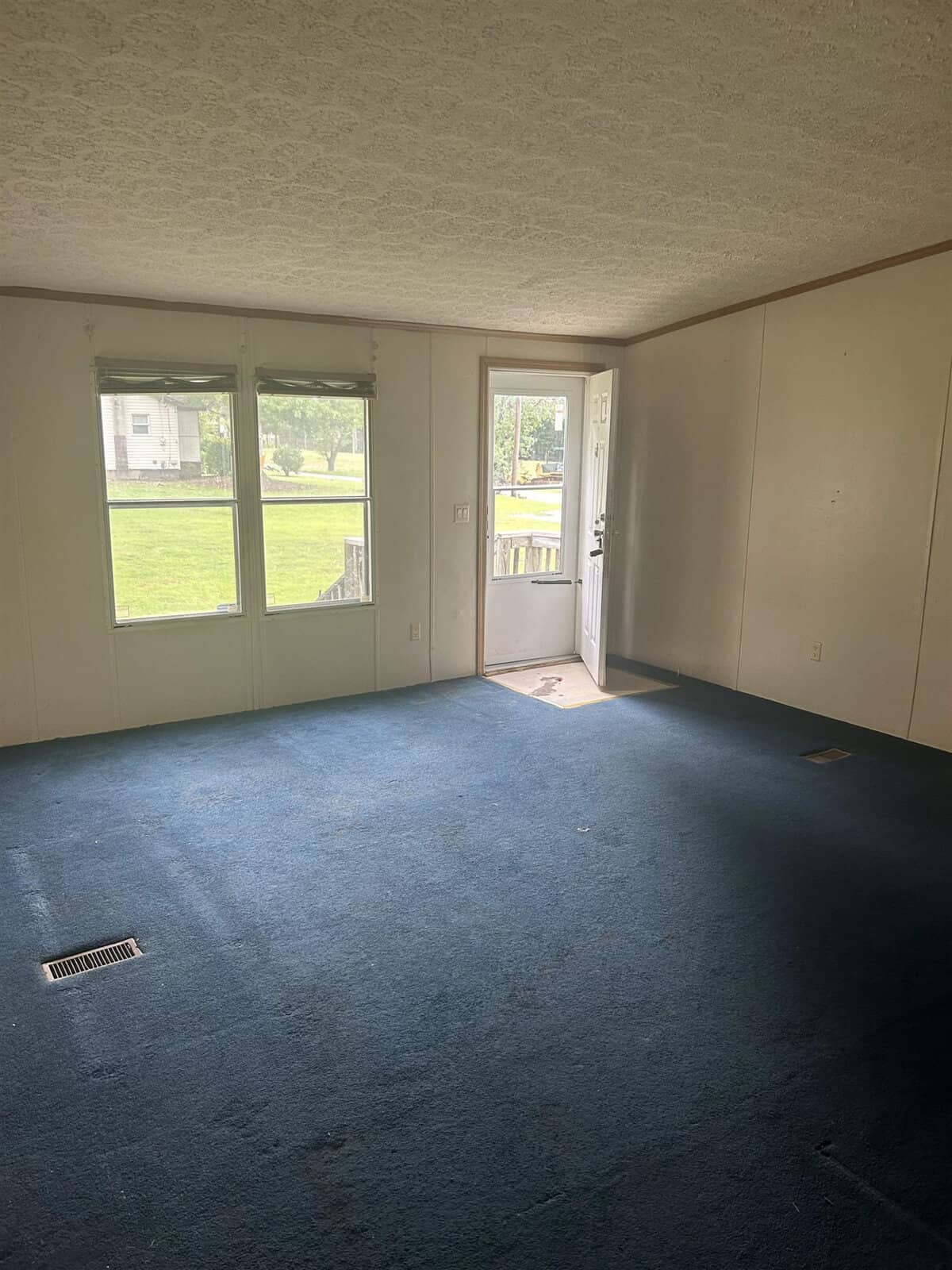 An empty room with blue carpet, two windows with blinds, and a door leading outside. Sunlight filters through the windows, illuminating part of the room. The walls are plain, and the ceiling has a textured pattern. A house is visible outside.
