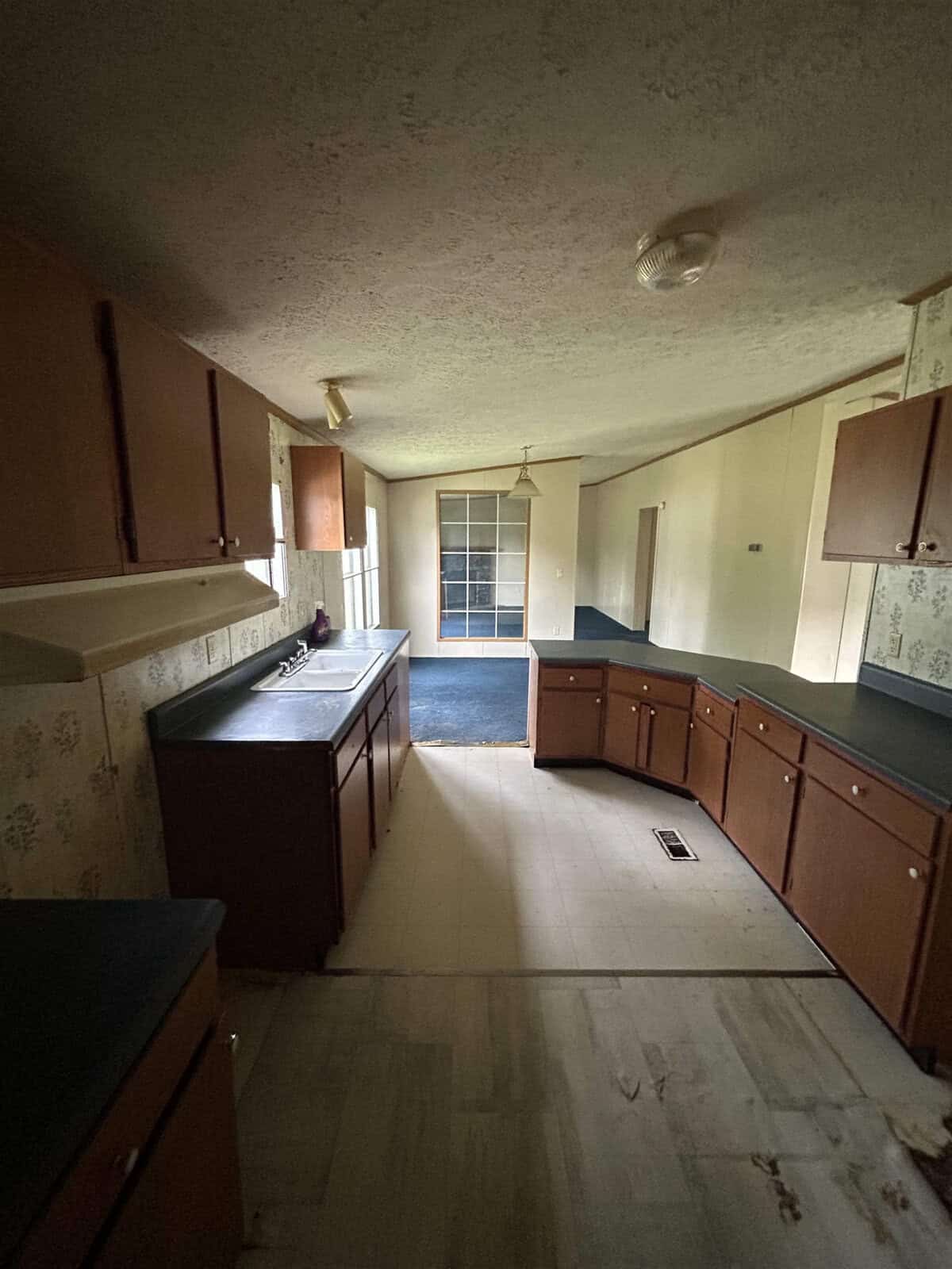 A dimly lit kitchen with wooden cabinets, dark countertops, and a double sink. A doorway leads to another room with a ceiling fan and large windows. The floor is a mix of worn vinyl and carpet. A bulb hangs from the ceiling.