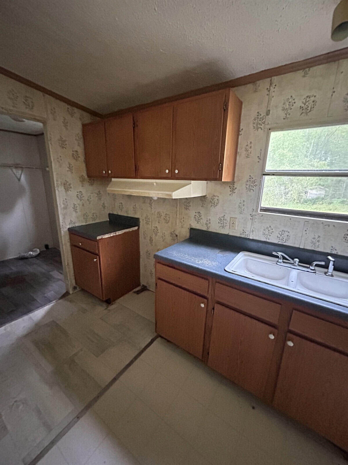 An empty kitchen with wooden cabinets, a white sink, and dark countertops. It's situated in a small space with floral wallpaper and a window to the right. There's an adjacent open area with visible flooring transitions.