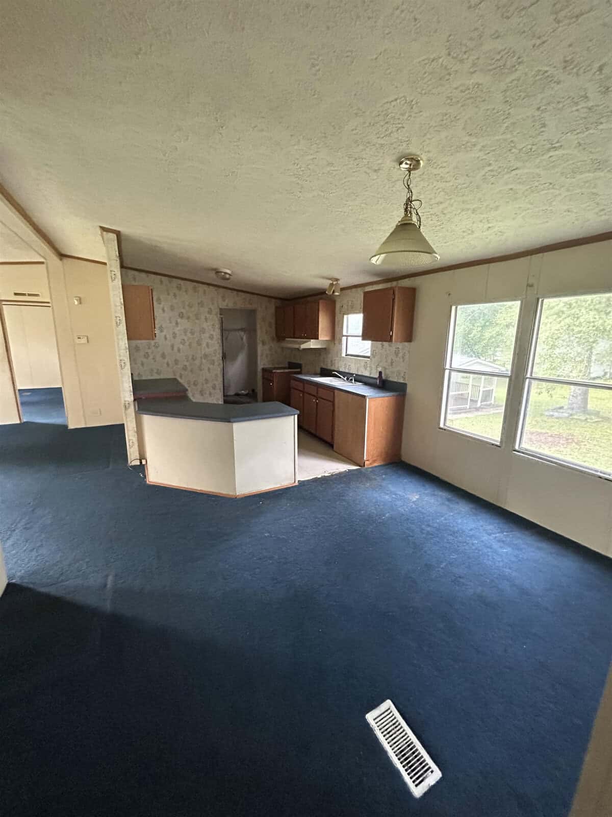 A room with blue carpet, featuring a small kitchen area with wooden cabinets, white countertops, and a hanging light fixture. Large windows provide natural light and a view of greenery outside. A hallway leads to other areas of the space.