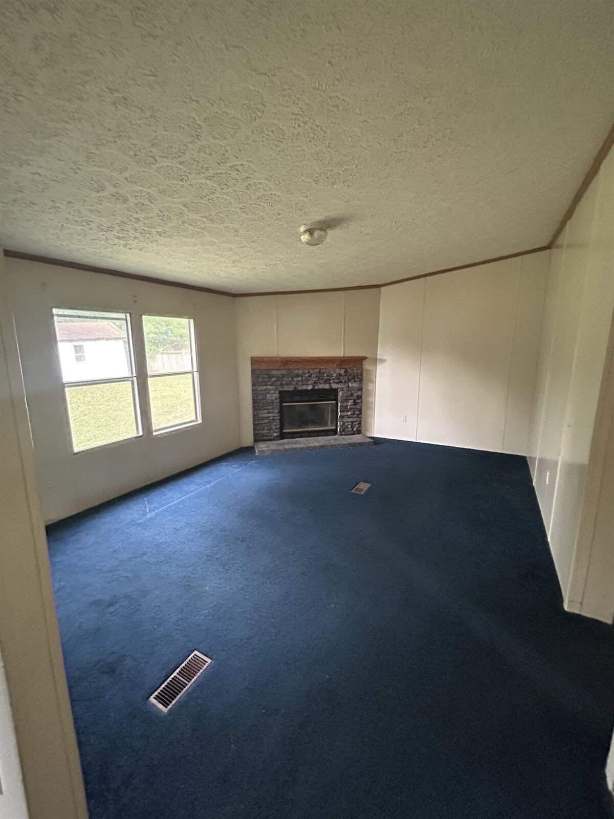 Room with blue carpet, white walls, and a textured ceiling. A stone fireplace with a wooden mantel is set against one wall. Three large windows provide natural light, and a ceiling light fixture is centered. There's a vent on the floor.