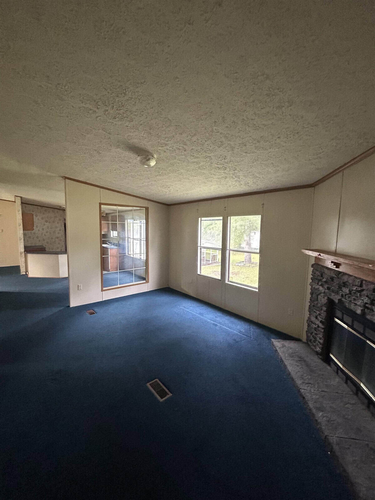 A vacant room with blue carpet, large windows, and a stone fireplace. The walls are light-colored, and a mirrored closet door reflects a portion of the room. The ceiling is textured, and there is natural light coming through the windows.