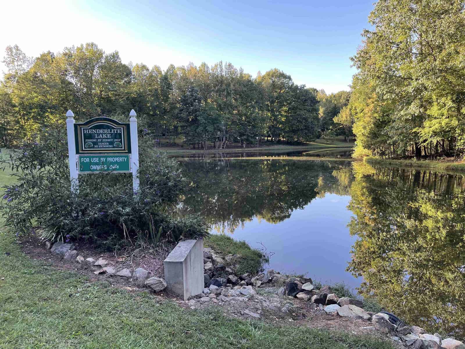 A tranquil pond surrounded by lush trees under a clear sky. A green and white sign reads "Hendersonbelte Lake, Private Property," with a smaller note about use for property owners. The foreground has grass and a few rocks near the water's edge.