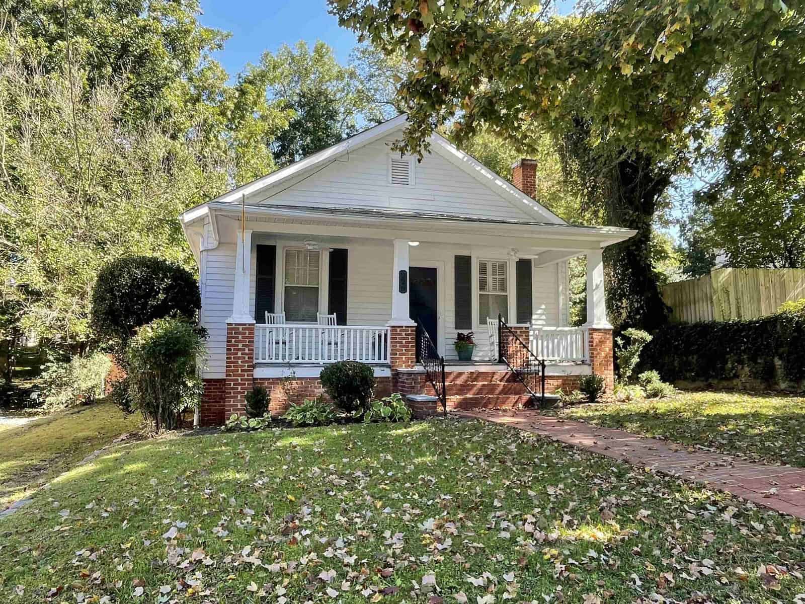 A quaint, white single-story house at 226 W Belle Street, Henderson NC, with a red brick pathway, flanked by trees and fallen leaves, on a sunny day.