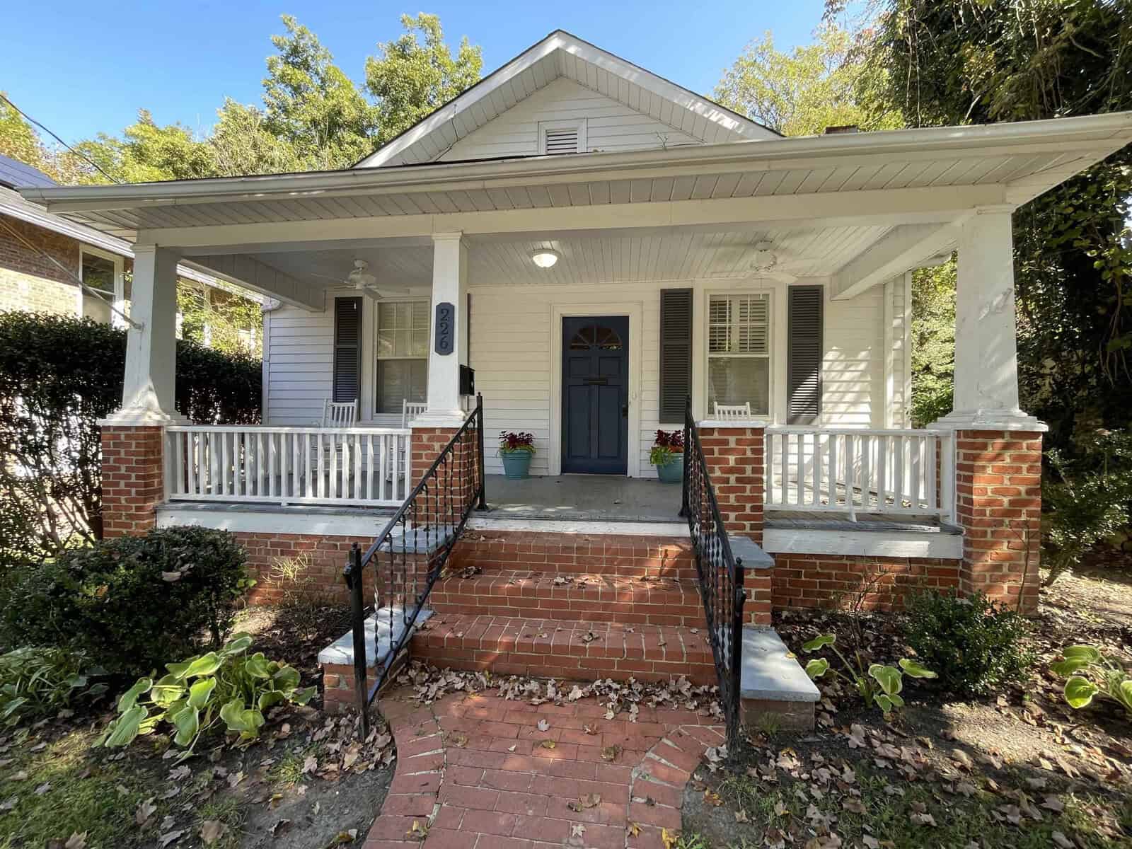 A single-story house with a covered front porch supported by columns, a brick staircase leading to a blue front door, flanked by two black shutters, and potted plants on each side.