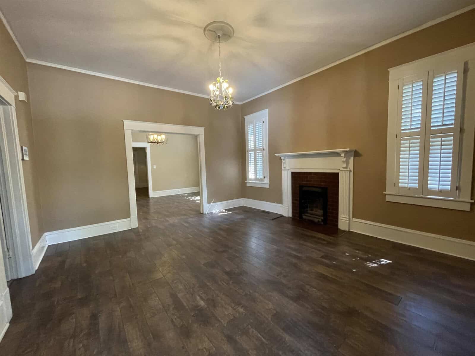 An empty room featuring polished hardwood floors, a fireplace, and windows with white shutters; two doorways lead to adjacent rooms.