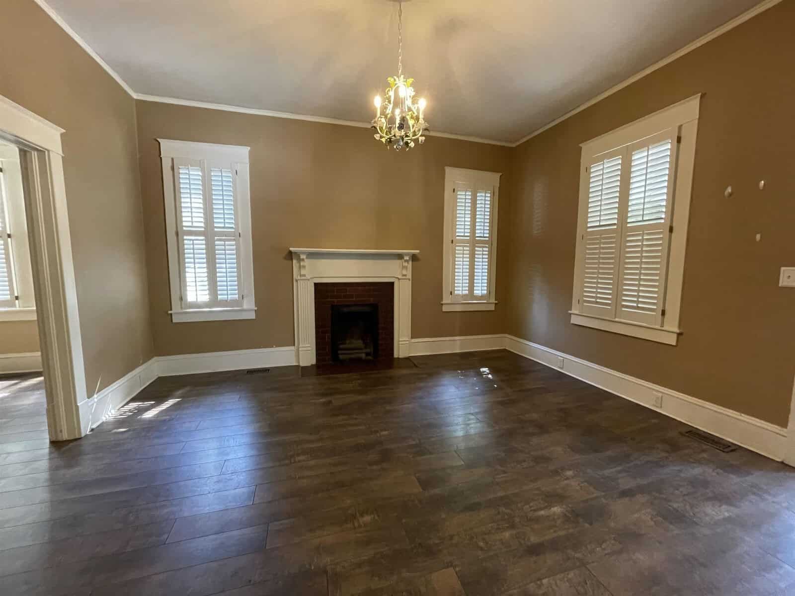 Empty room with beige walls, dark hardwood flooring, a white fireplace, closed white shutters on windows, and a hanging chandelier.