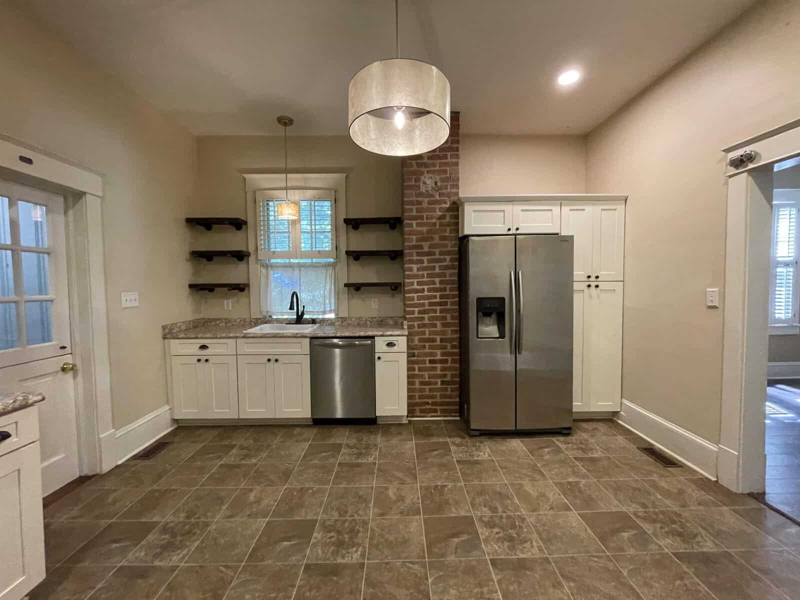 A well-lit kitchen with tiled flooring, featuring stainless steel appliances, white cabinetry, and a brick accent wall with open shelves.