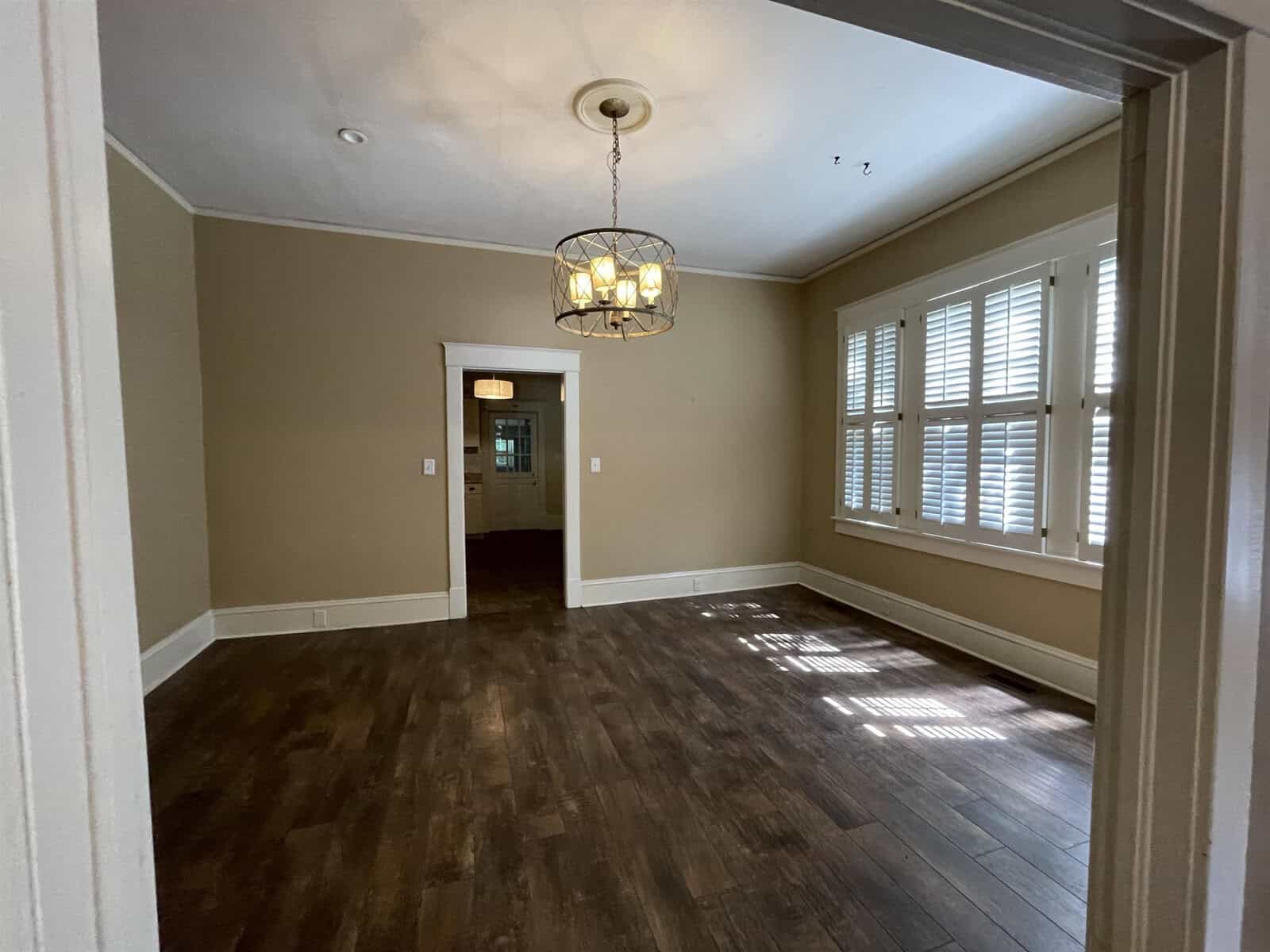 An empty room with hardwood floors, beige walls, a chandelier, and large windows allowing natural light to cast shadows on the floor.