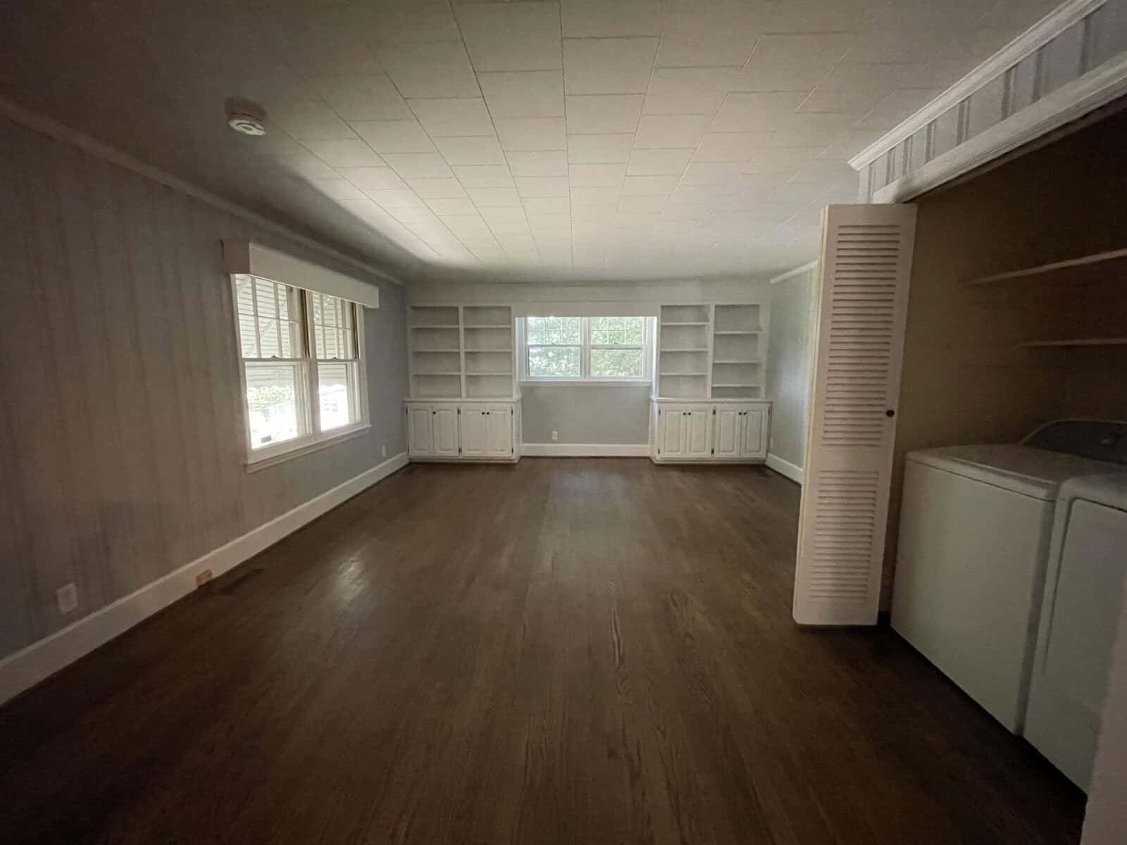 An empty room with wooden flooring, white walls, built-in shelving units, and a washing machine at the side.
