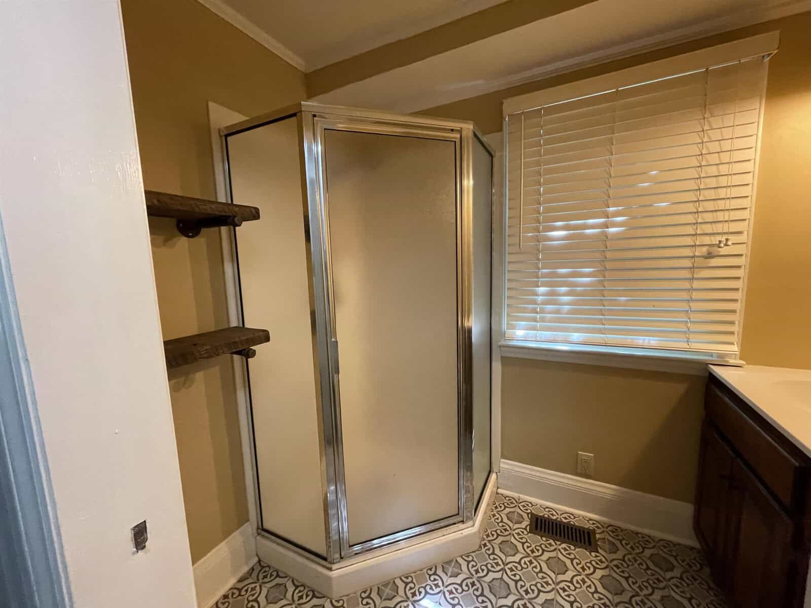A corner shower with sliding doors installed in a room with beige walls, complemented by a patterned floor and adjacent to a window with horizontal blinds. to the left, two wooden shelves are affixed to the wall.