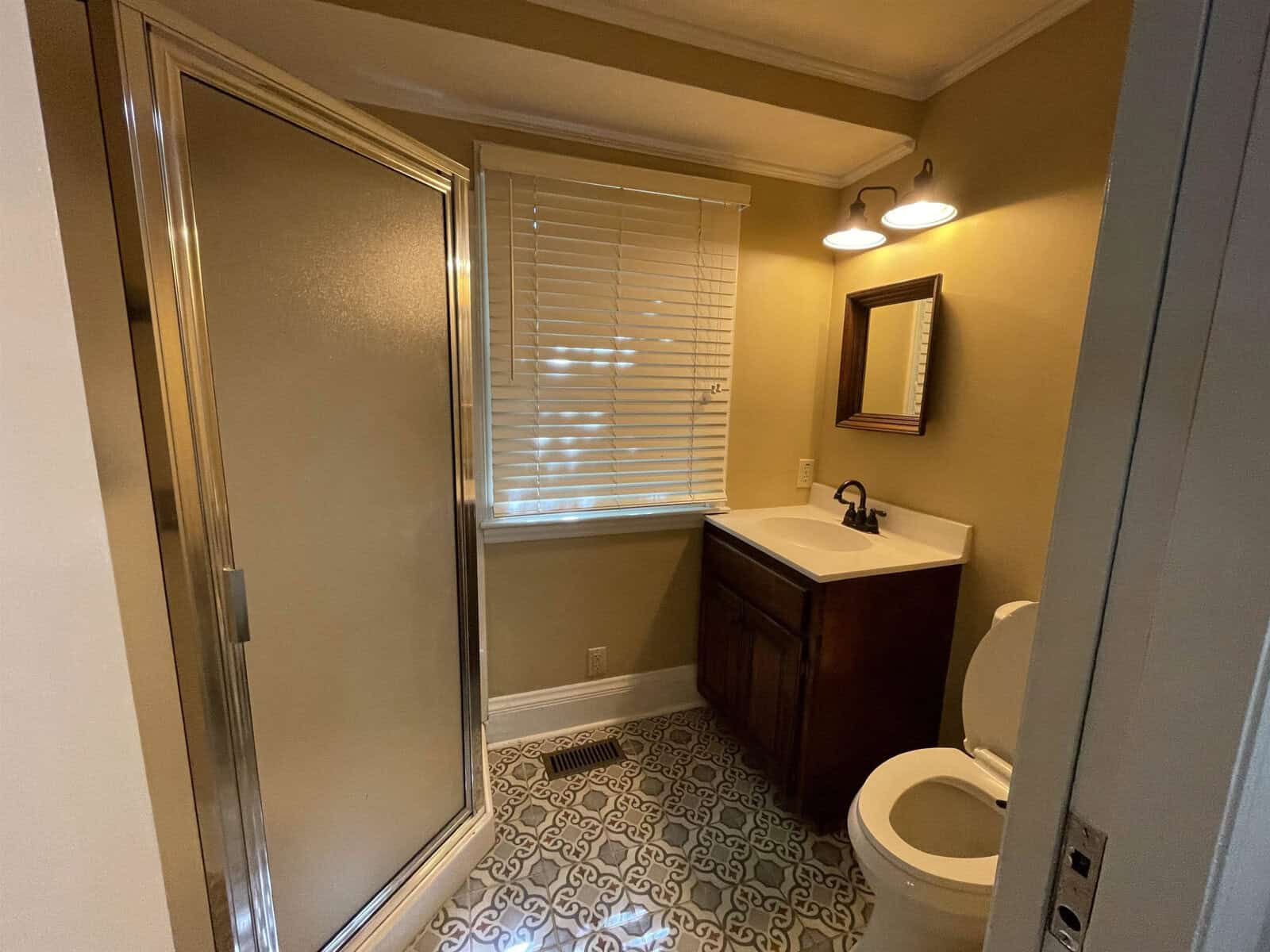 A small, neatly kept bathroom with a shower enclosure on the left, a toilet, and a sink with a cabinet underneath. the room has patterned flooring, a window with blinds, and a framed picture on the wall.